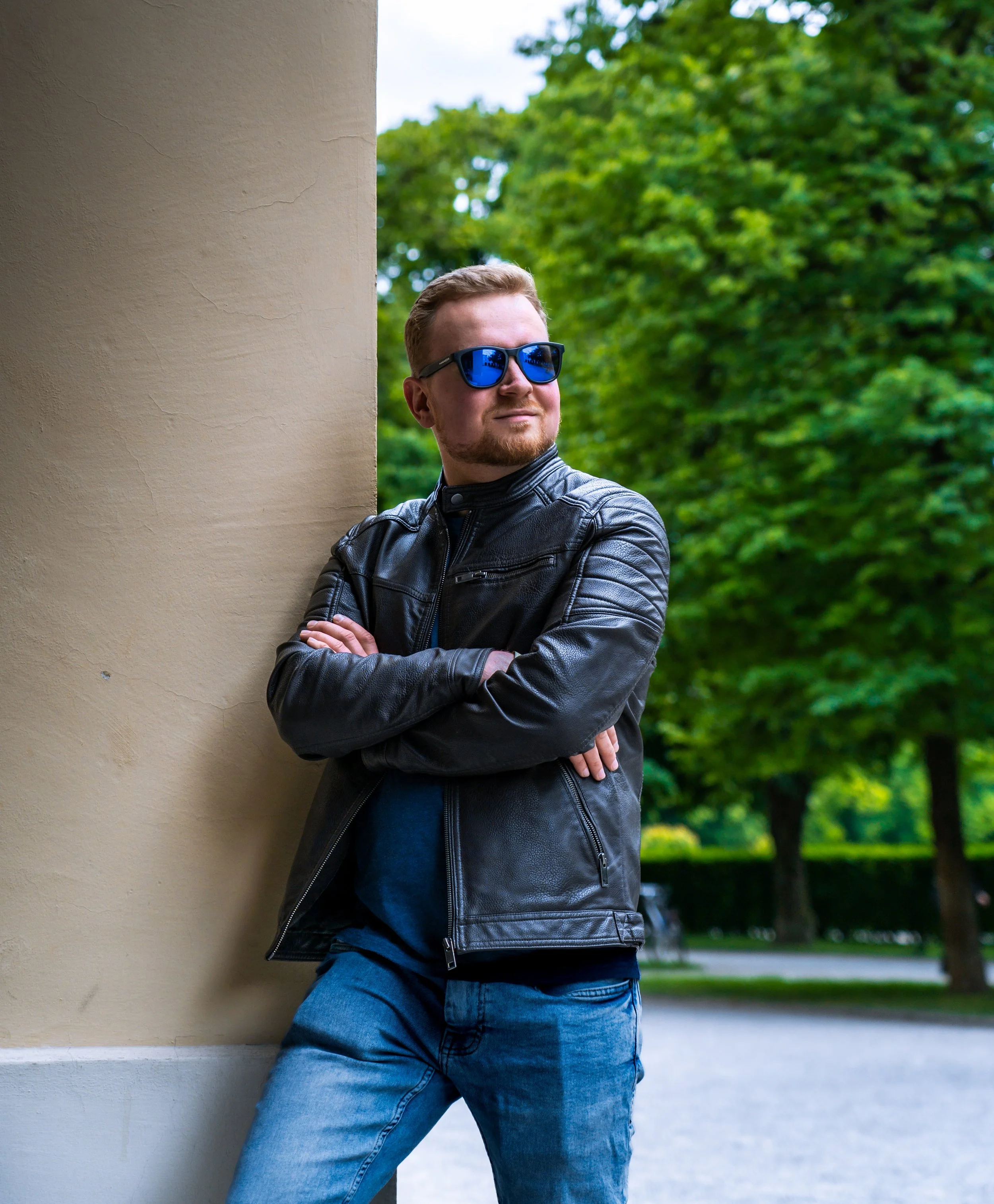 Junge Mann trägt schwarze Lederjacke und blaue Sonnenbrille, lehnt an einer Wand, umgeben von grünen Bäumen im Park.