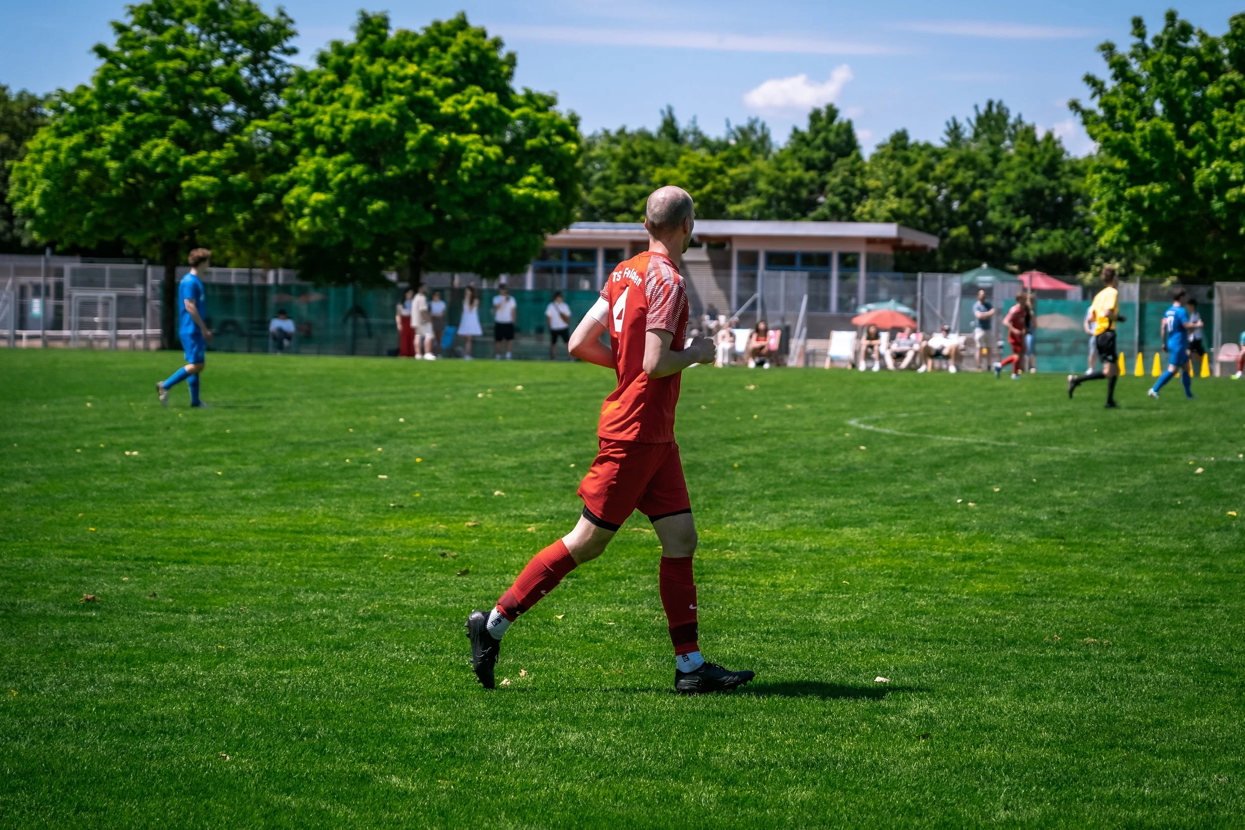 Mann in rotem Fußballtrikot läuft auf einem grünen Fußballplatz, im Hintergrund Zuschauer und Bäume, sonniger Tag.