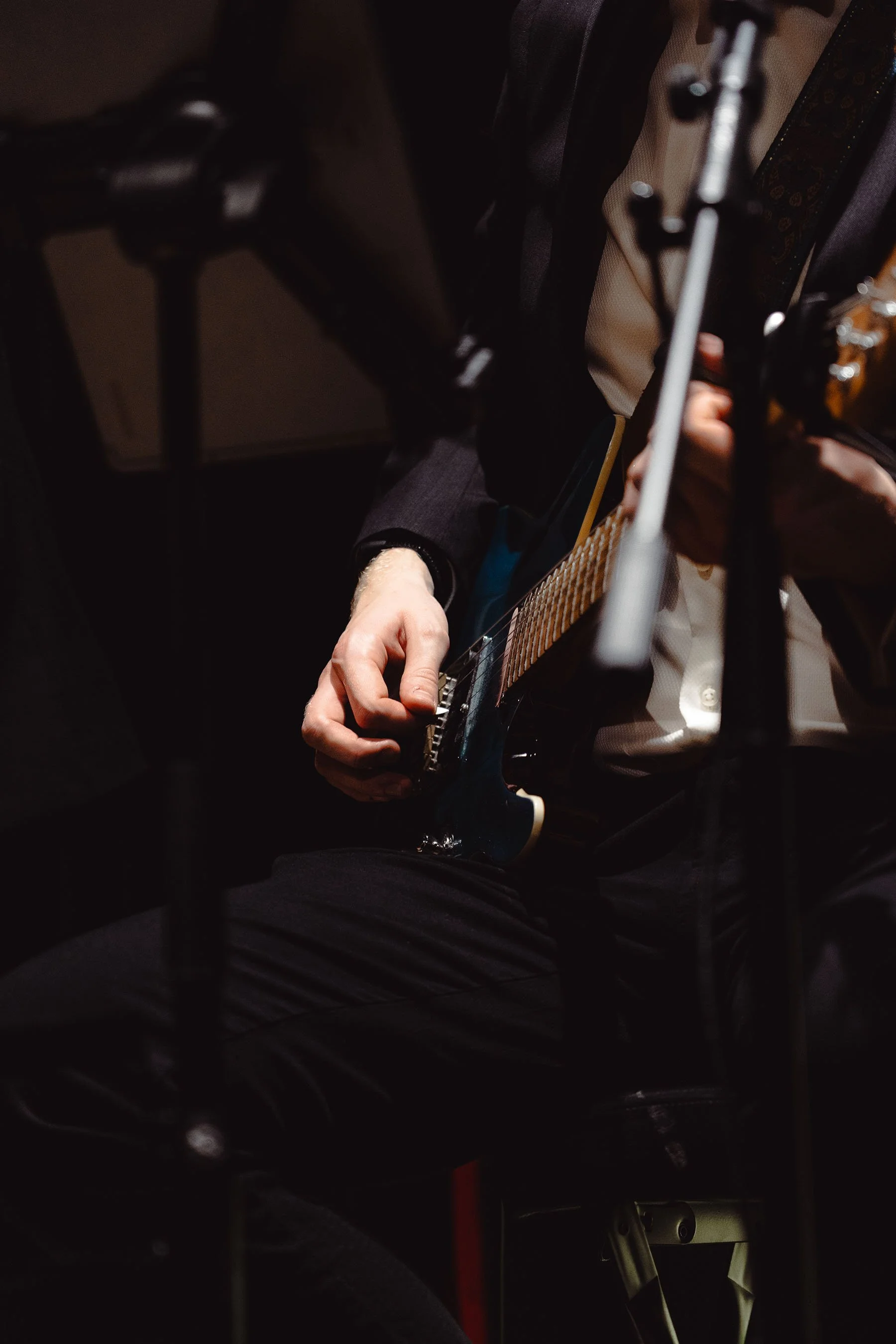 Eine Person spielt Gitarre, teilweise verborgen durch dunkle Objekte im Vordergrund, auf einer Bühne.