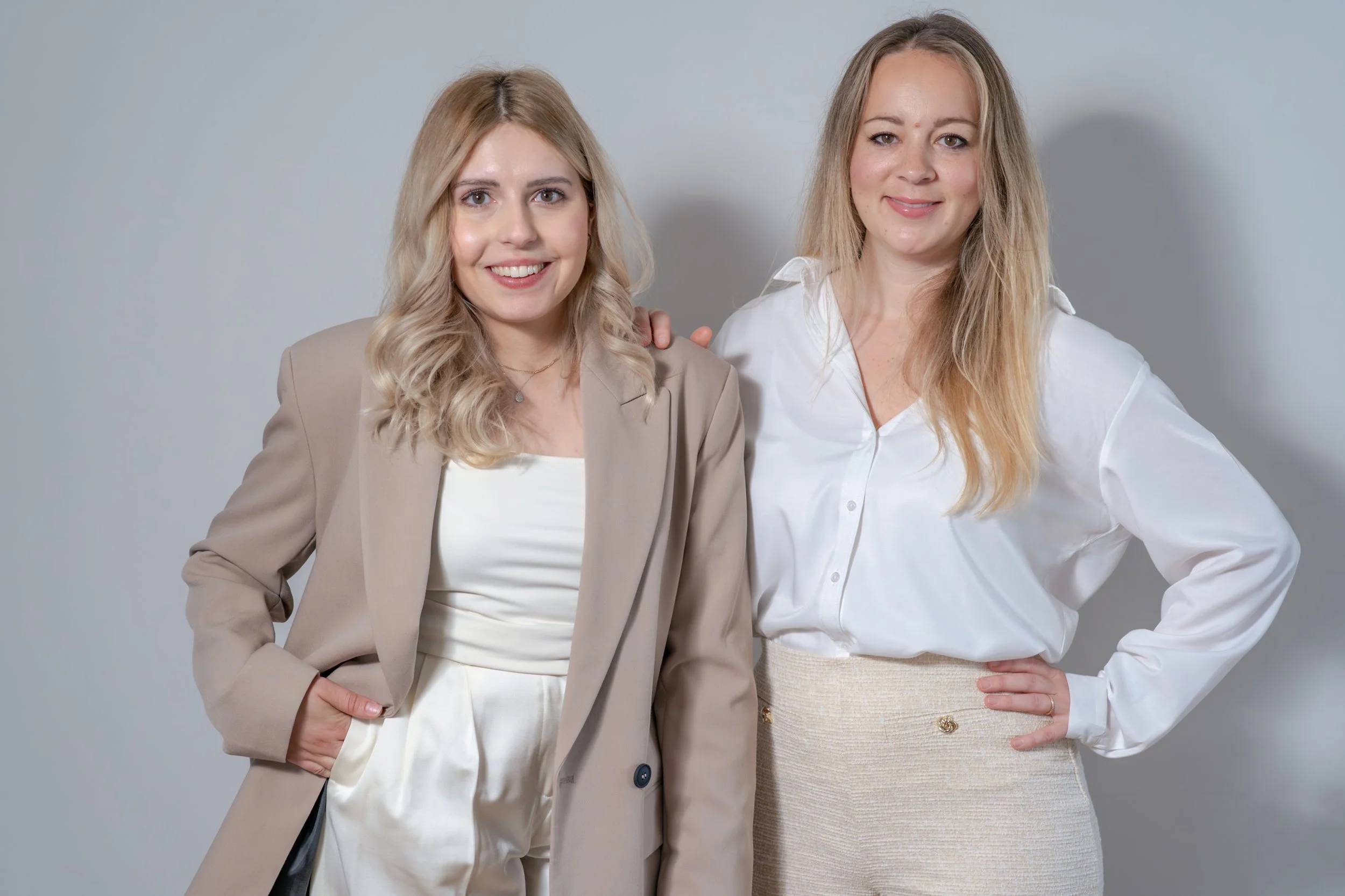 Zwei lächelnde Frauen stehen nebeneinander vor einem neutral grauen Hintergrund, eine trägt einen beige Business-Anzug, die andere ein weißes Hemd mit cremefarbener Textur
