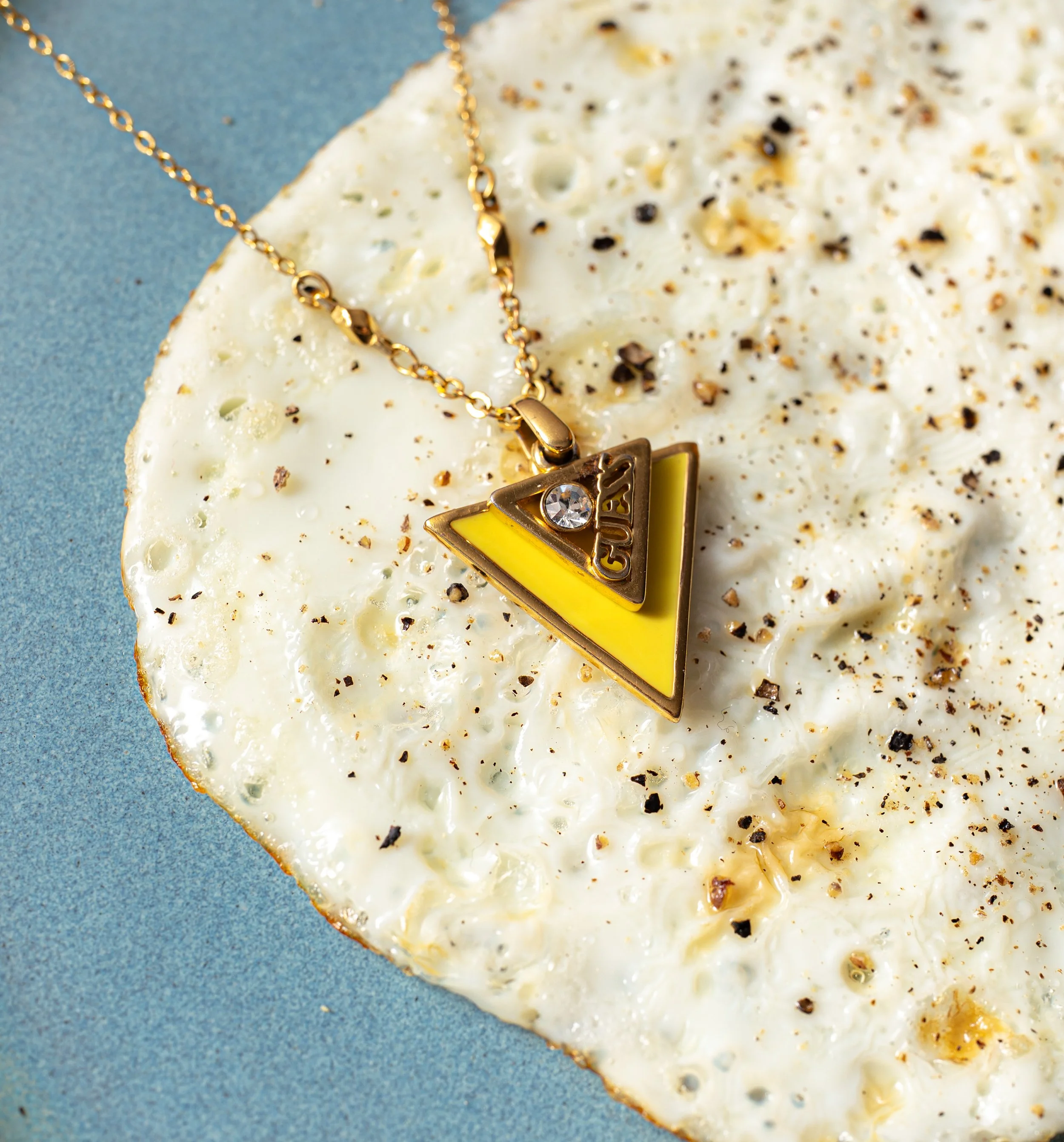 Goldene Halskette mit einem Anhänger auf einer weißen, mit schwarzen Pfefferkörnern und Kräutern bestreuten Käseplatte