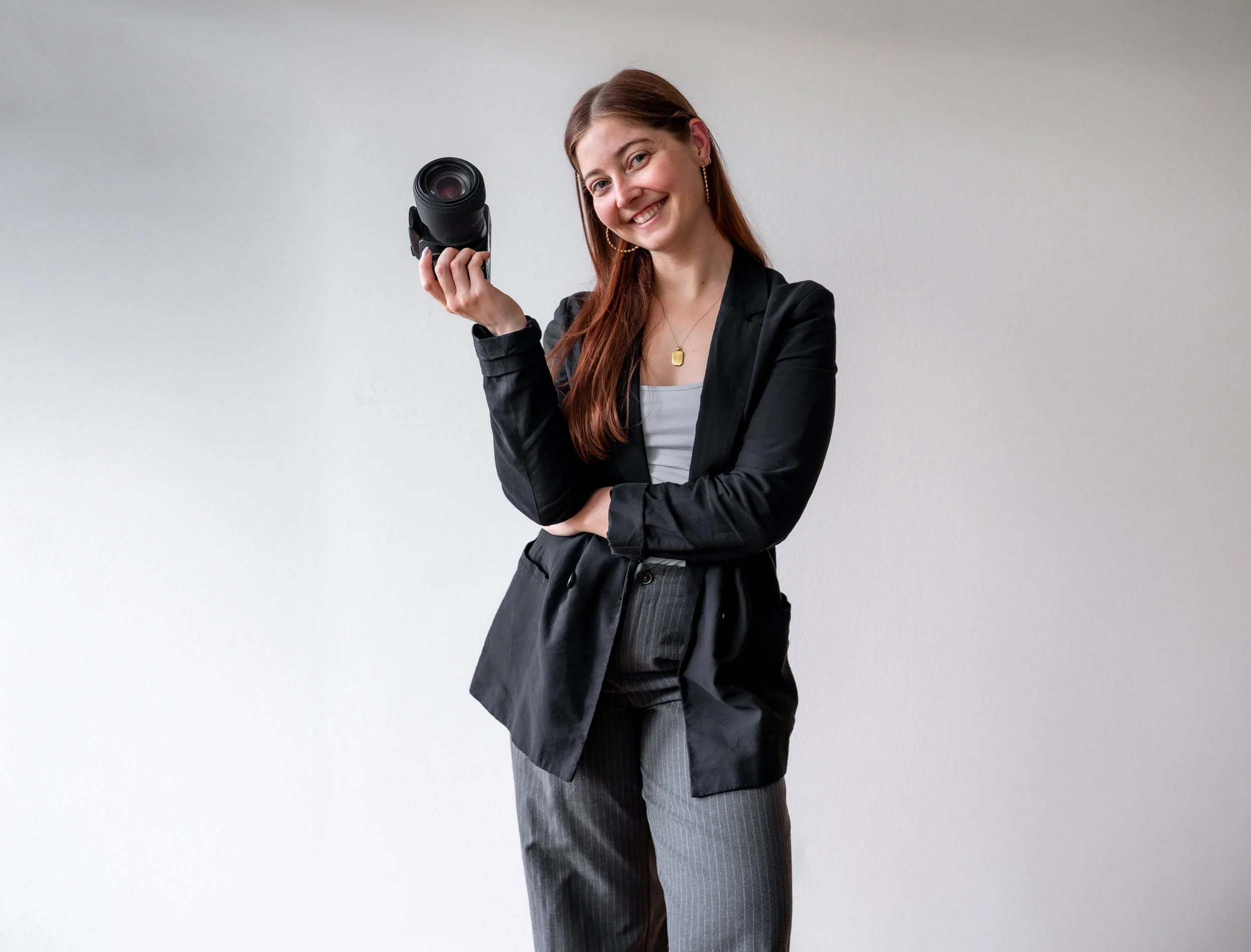 Eine lachende junge Frau mit braunen Haaren trägt eine schwarze Jacke und graue Streifenhose, hält eine Digitalkamera in der Hand und steht vor einer weißen Wand.