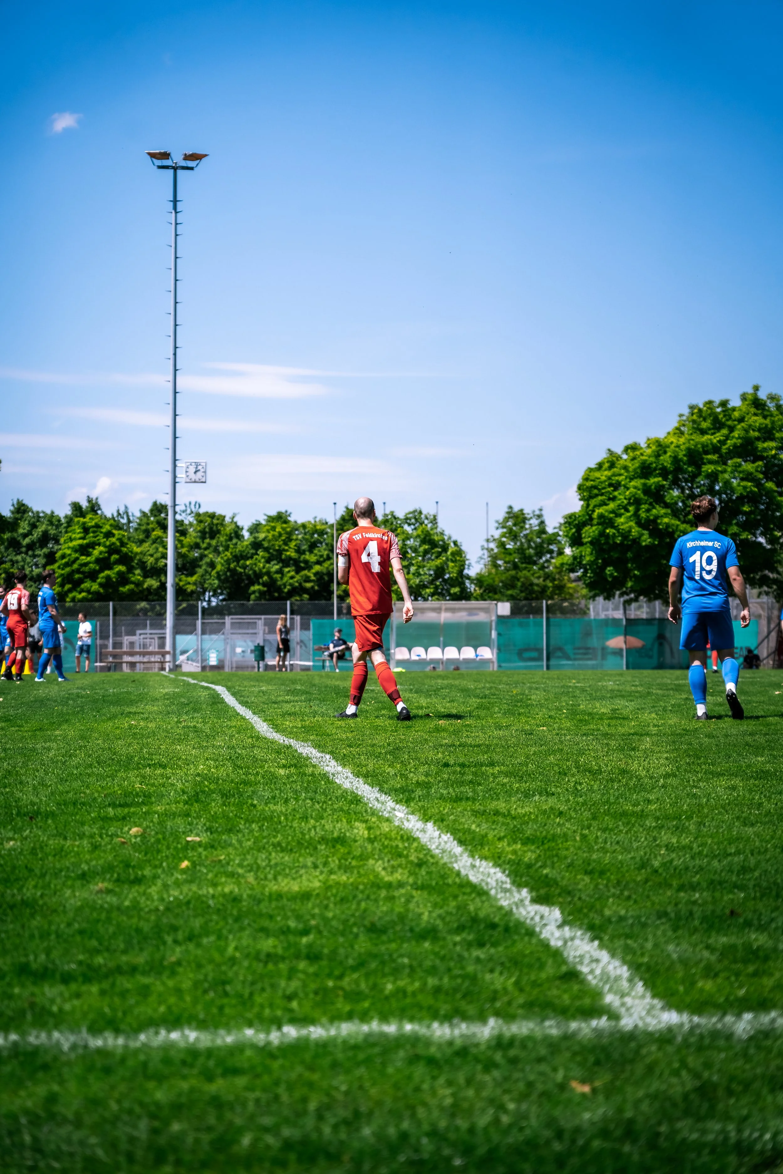 Fußballspieler auf einem grünen Spielfeld bei sonnigem Wetter, hinter Bäume und ein blauer Himmel.
