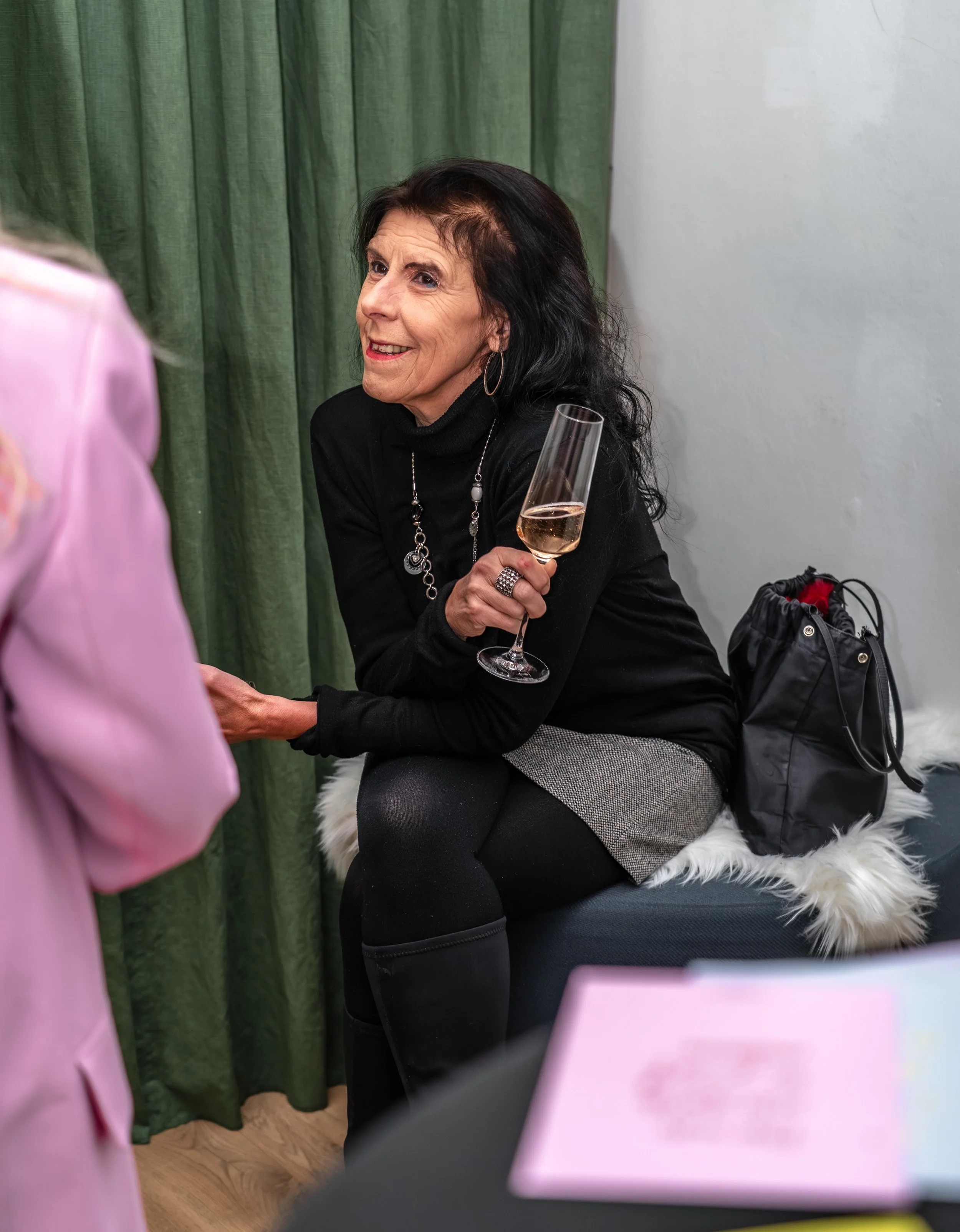 Eine ältere Frau mit schwarzen Haaren, die ein Glas Sekt hält, sitzt auf einem Sofa und unterhält sich mit jemandem, der nur teilweise im Bild zu sehen ist, vor einem grünen Vorhang und einer weißen Wand.