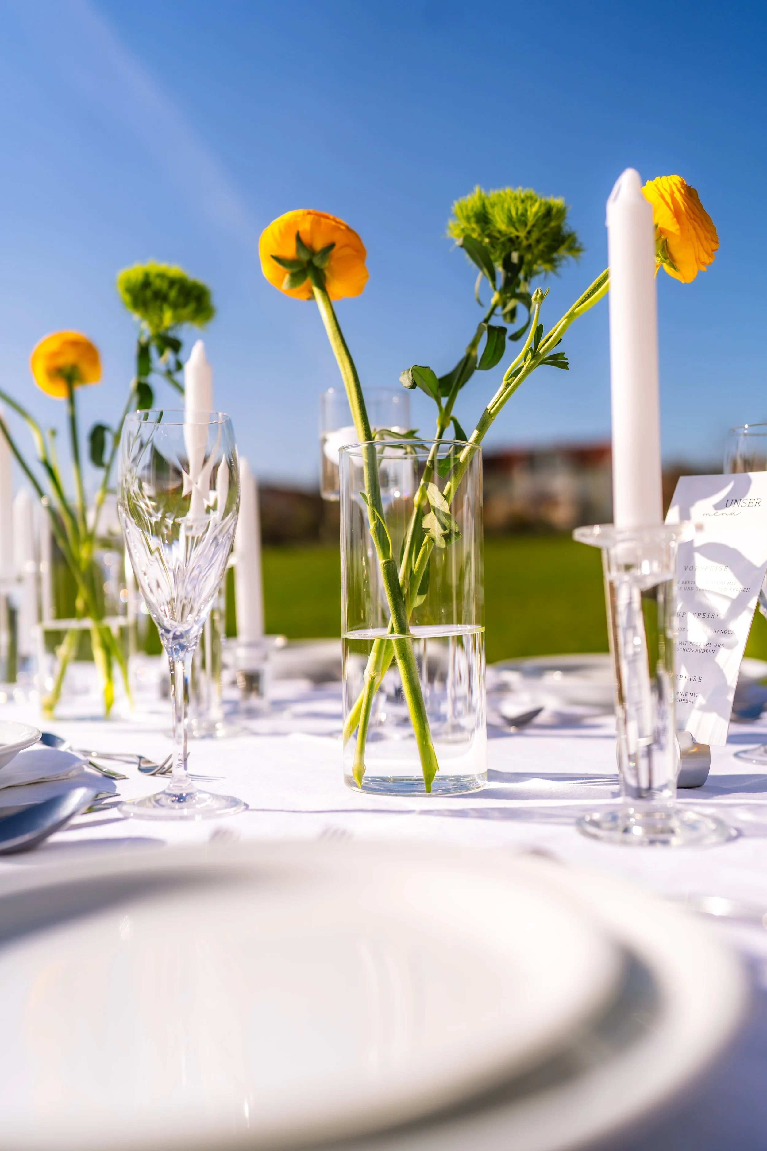 Blumenstrauß mit gelben und grünen Blumen in einer Glaskaraffe, umgeben von Kerzen und Gläsern auf einem weißen Tisch bei sonnigem Himmel.