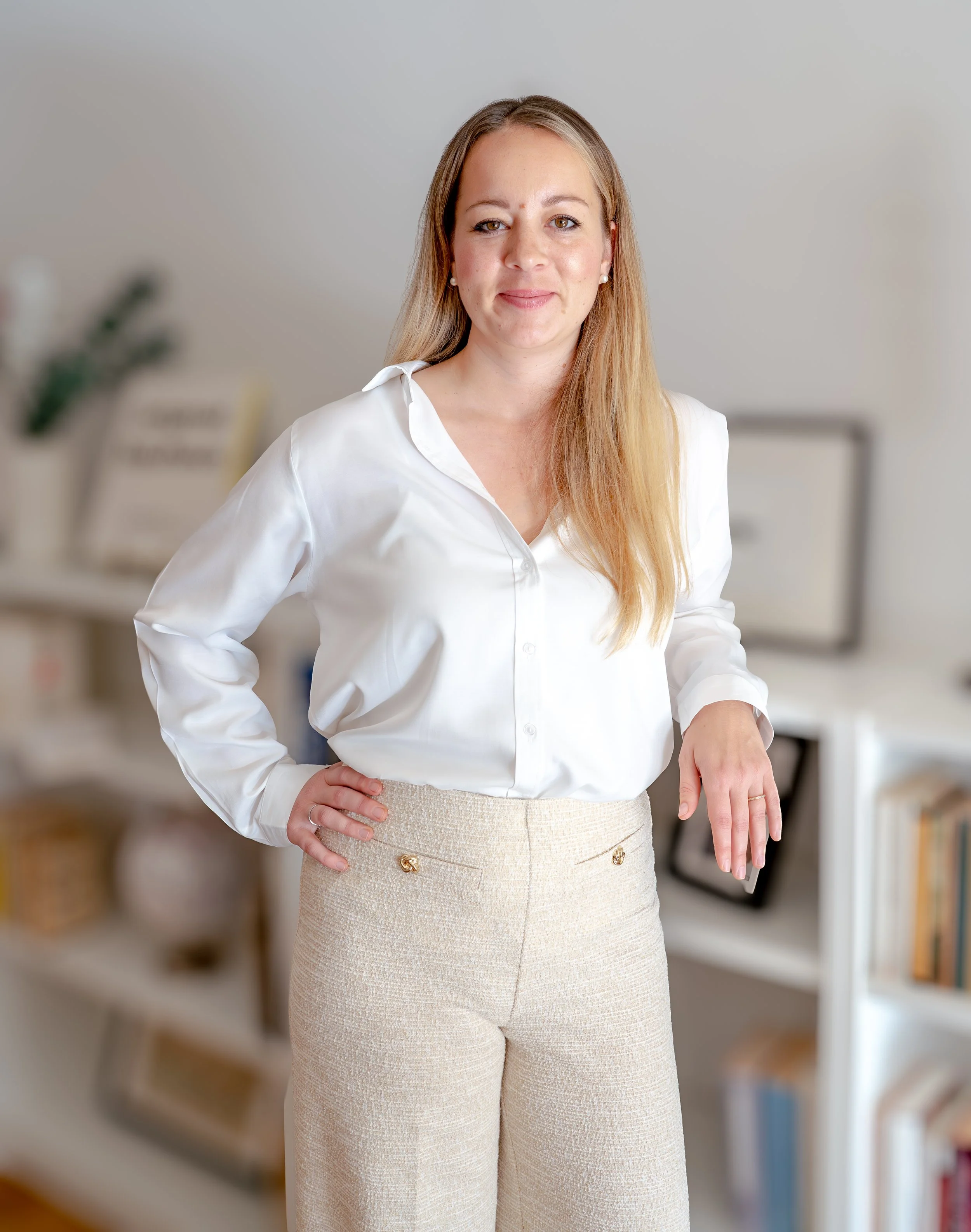 Eine Frau mit langen, blonden Haaren trägt eine weiße Bluse und beigefarbene Hosen und steht in einem modernen Büroraum.