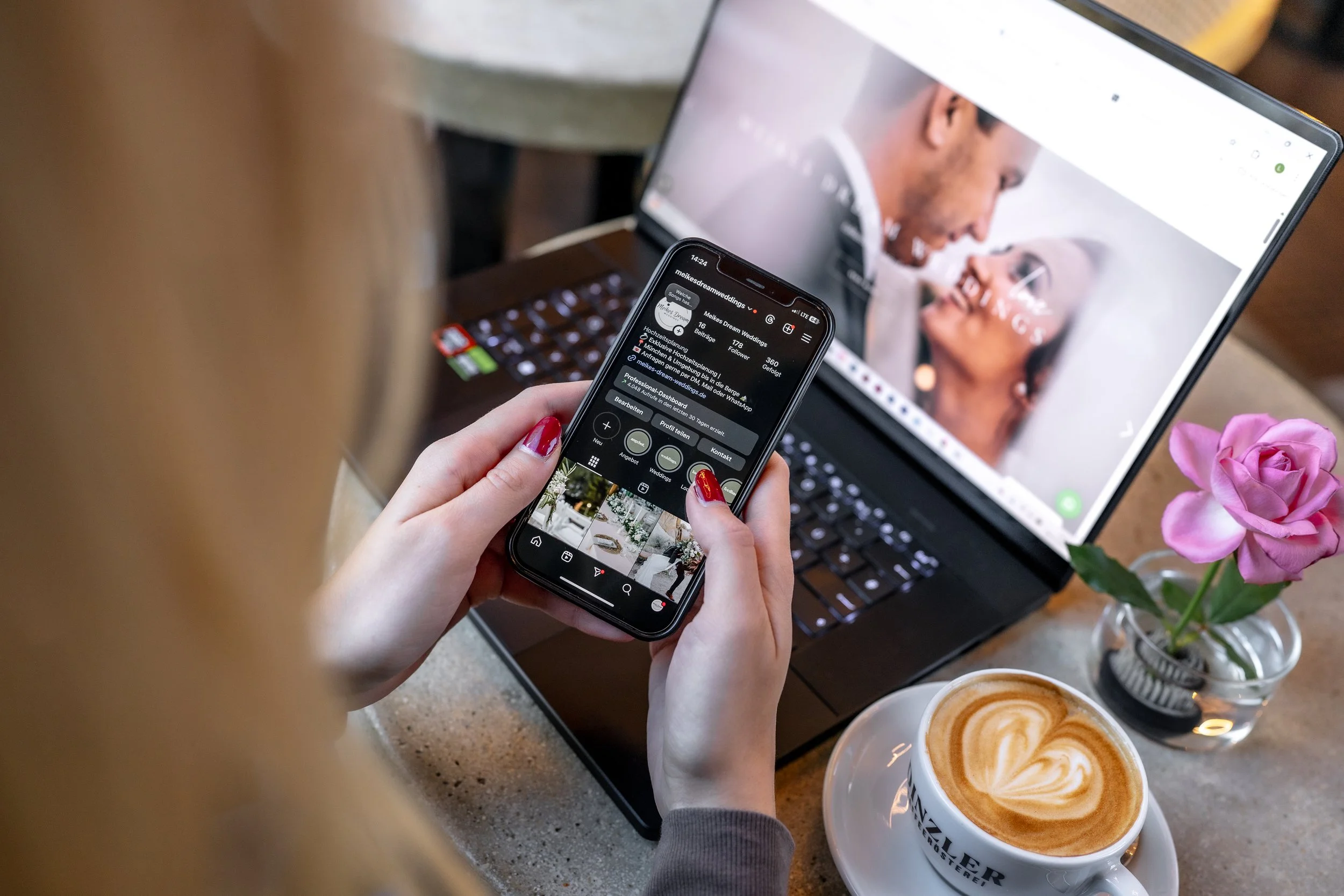 Eine Person hält ein Smartphone, das einen Instagram-Account mit Hochzeitsfotos zeigt, vor einem Laptop mit einem Foto eines lachenden Paares. Auf dem Tisch stehen zwei Cappuccinos mit Herzmuster, eine Vase mit pinken Rosen und eine Tasse.