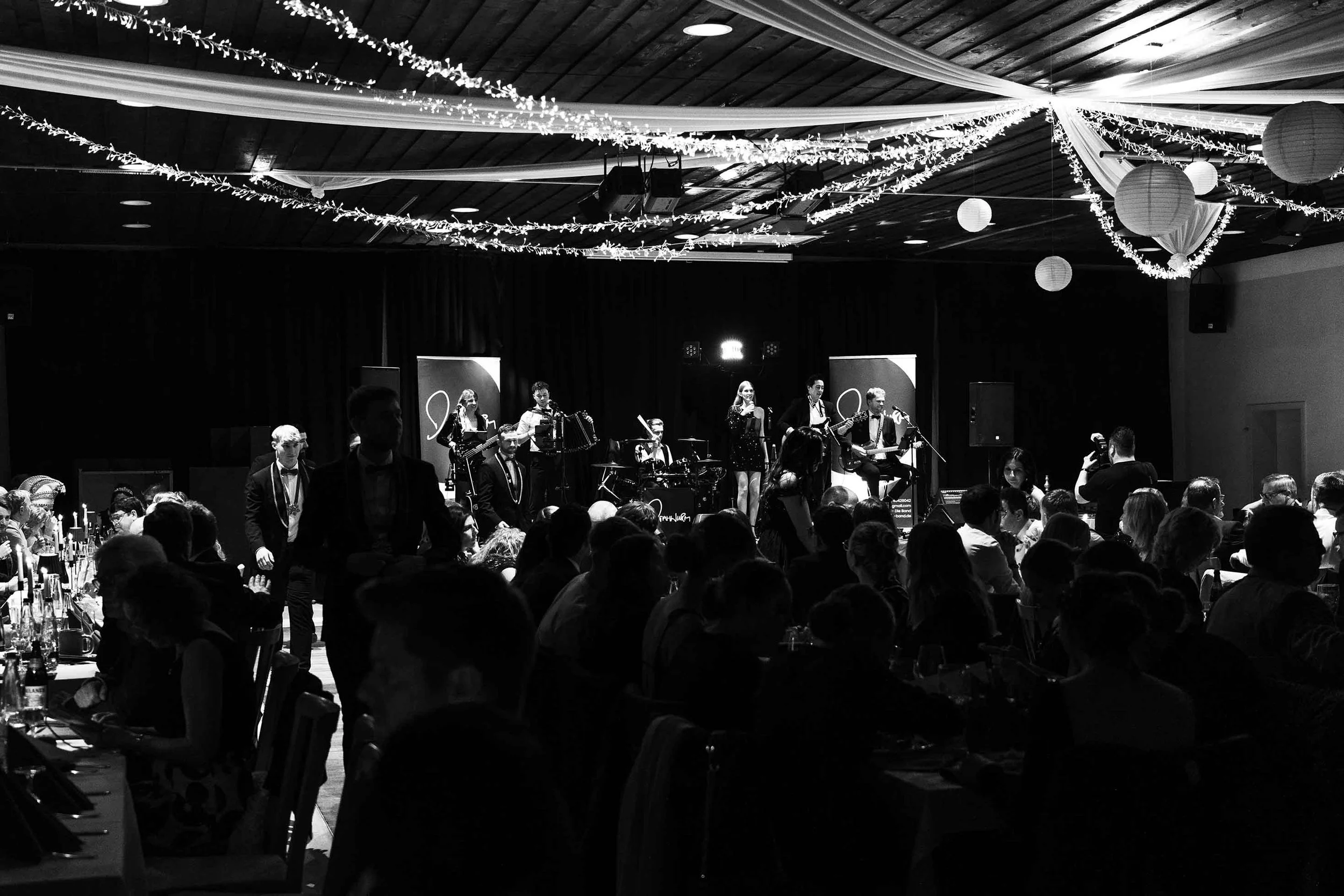 Eine schwarz-weiße Aufnahme einer festlich dekorierten Veranstaltung mit einer Bühne, auf der eine Band spielt. Das Publikum sitzt an Tischen, die mit Kerzen und Flaschen beleuchtet sind. Der Raum ist mit Lichterketten und Papierlaternen geschmückt.