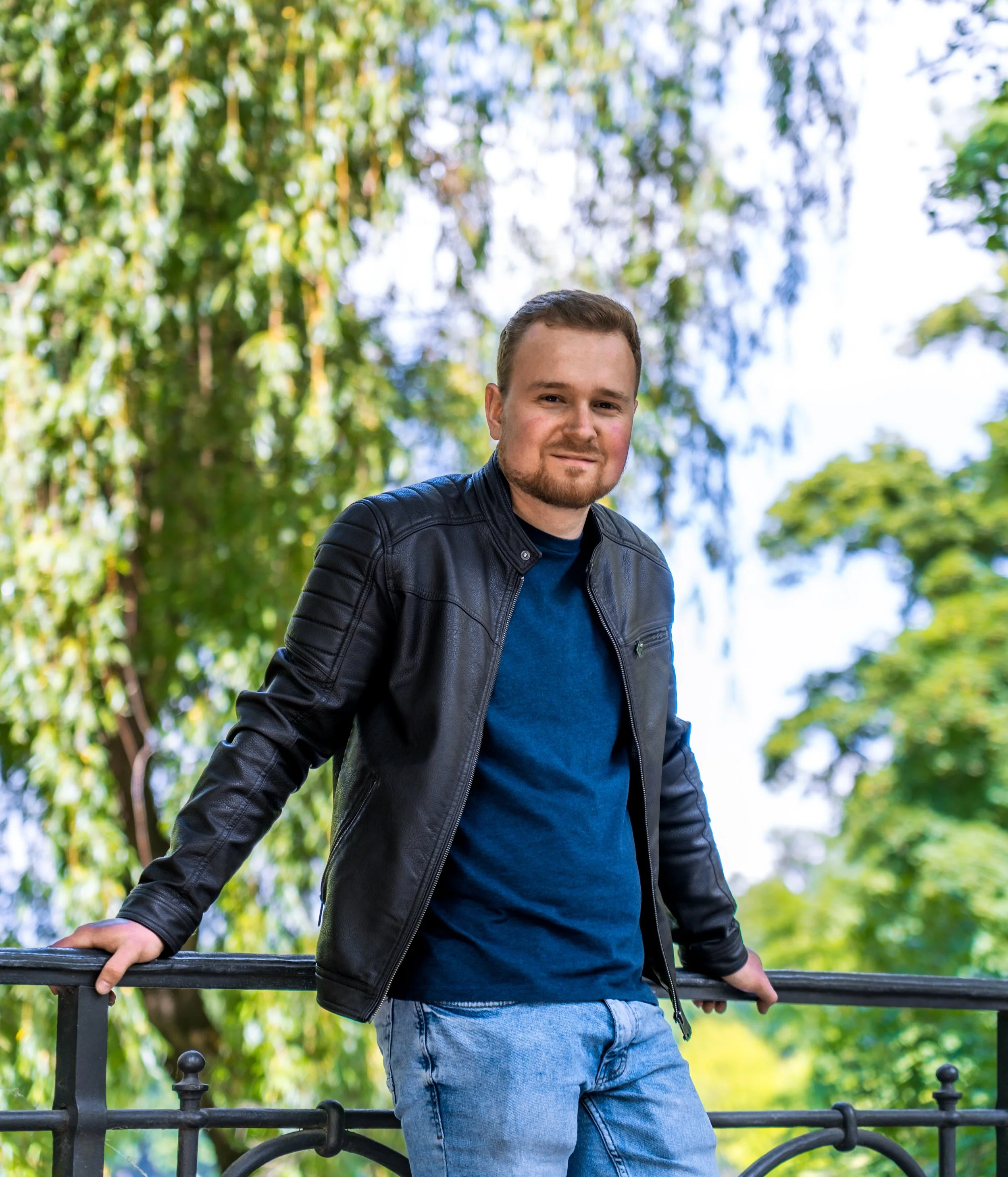 Ein junger Mann steht an einem Zaun im Freien, umgeben von grünen Bäumen und Himmel, trägt eine dunkle Lederjacke, einen blauen Pullover und Jeans, und lächelt leicht.