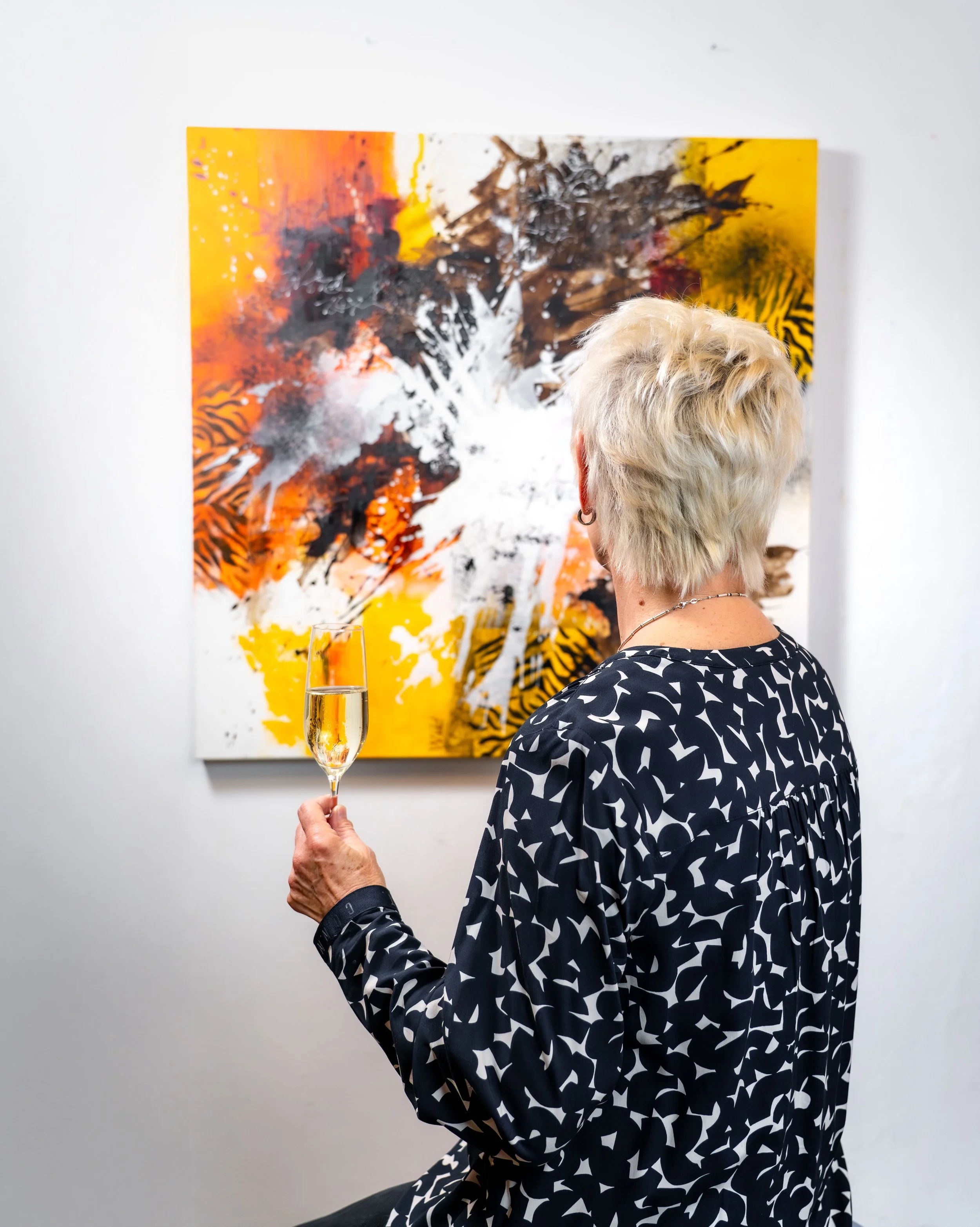 Eine Frau mit kurzen, blonden Haaren betrachtet ein abstraktes Gemälde in einer Galerie, während sie ein Glas Sekt hält.