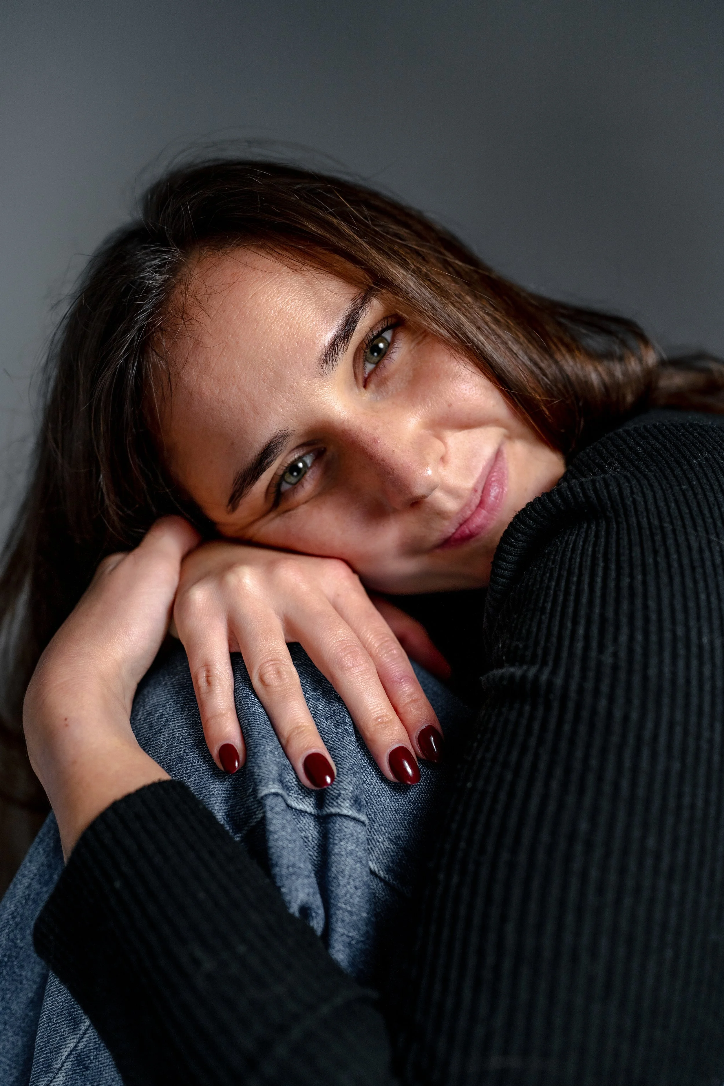Nahaufnahme eines lächelnden jungen Frauen mit braunen Haaren, die auf ihrer Seite liegt, die Hand unter dem Kopf, trägt dunklen Pullover und Jeans, roter Nagellack, schaut direkt in die Kamera.