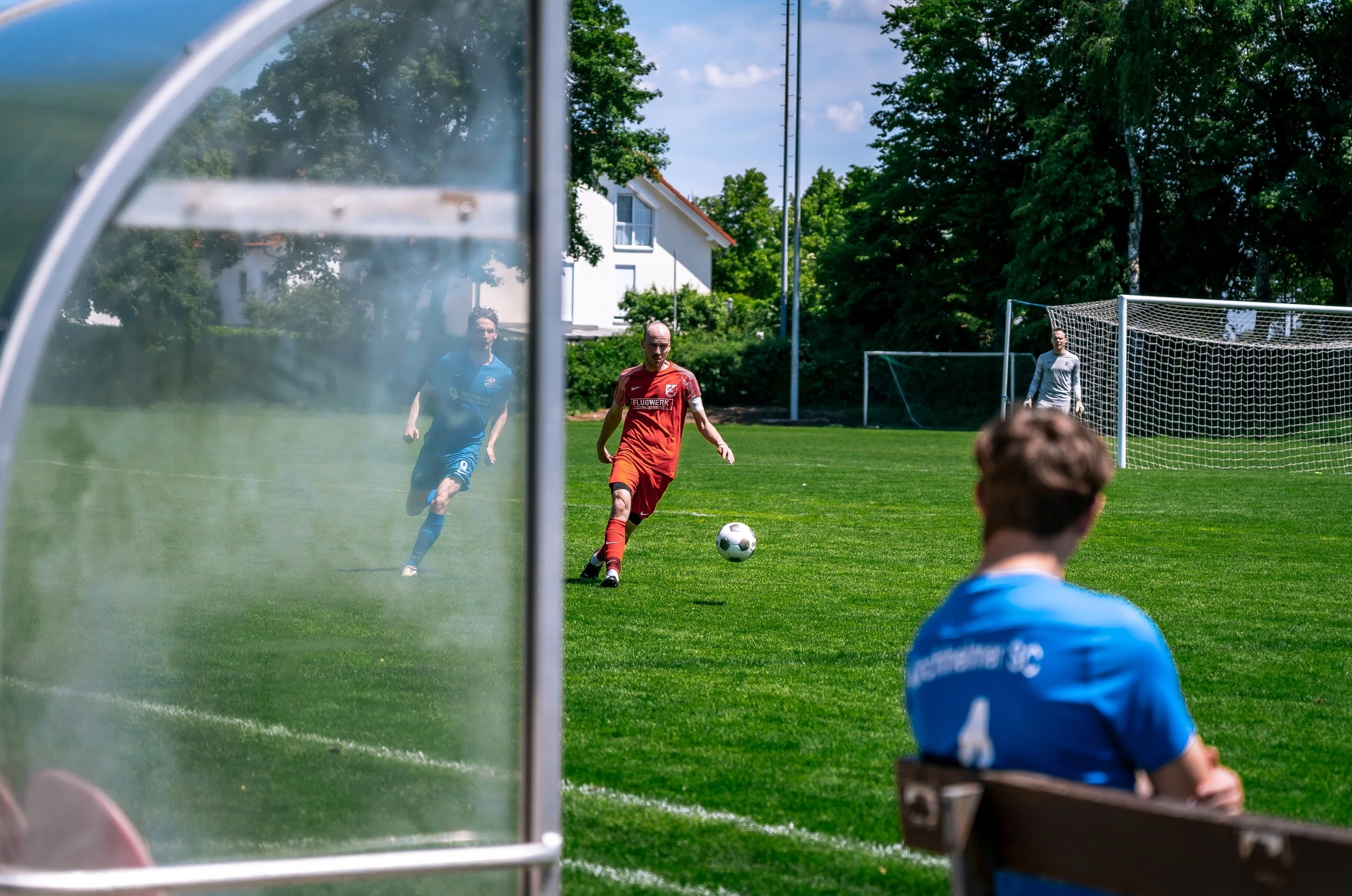 Fußballspiel auf einem grünen Feld, mit Spielern, Schiedsrichter, Tor und Zuschauern im Hintergrund, fotografiert durch eine Glasvitrine.
