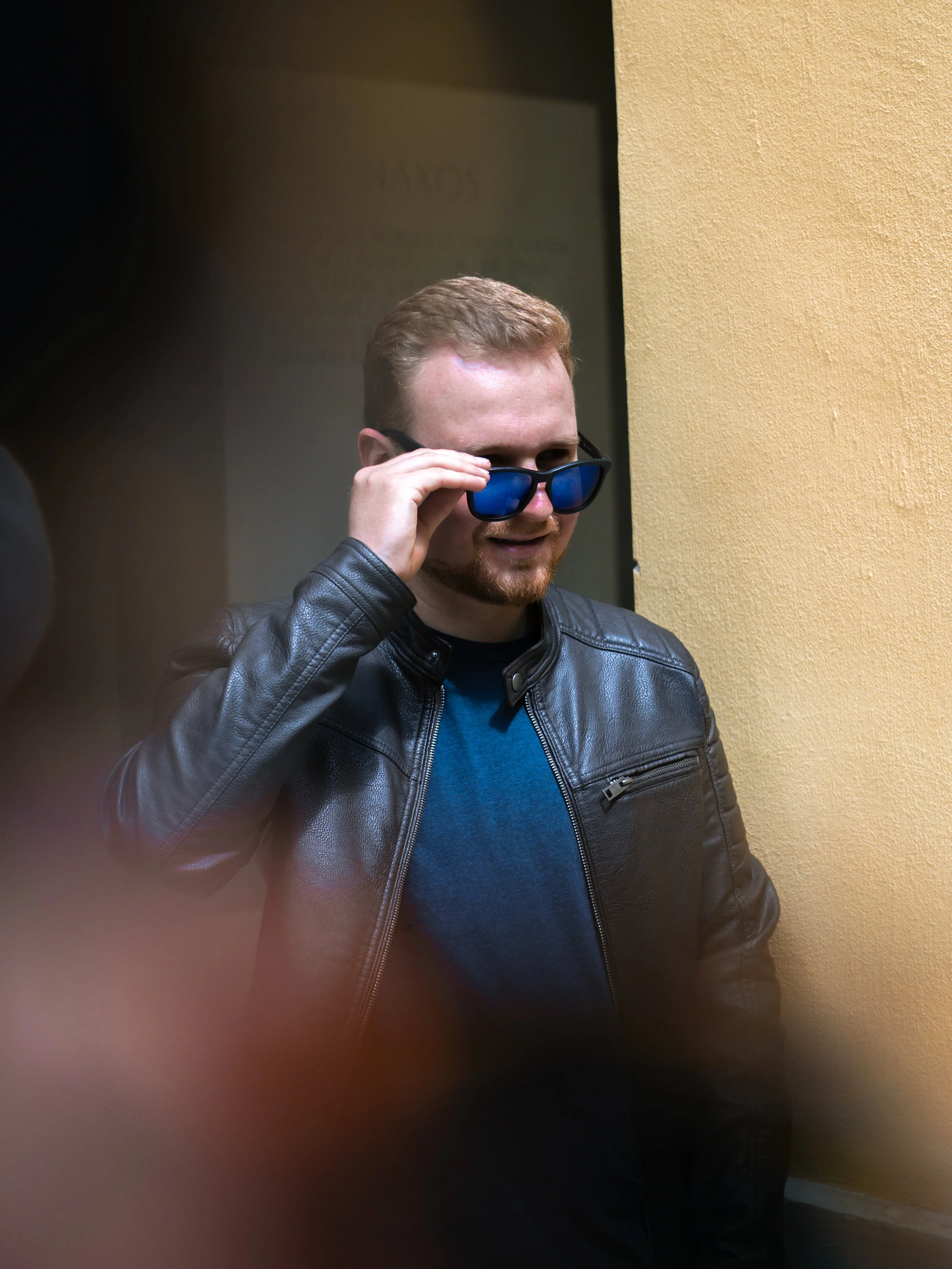 Ein Mann mit roten Haaren und Bart trägt eine schwarze Lederjacke und eine blaue Brille. Er steht vor einer hellgelben Wand.