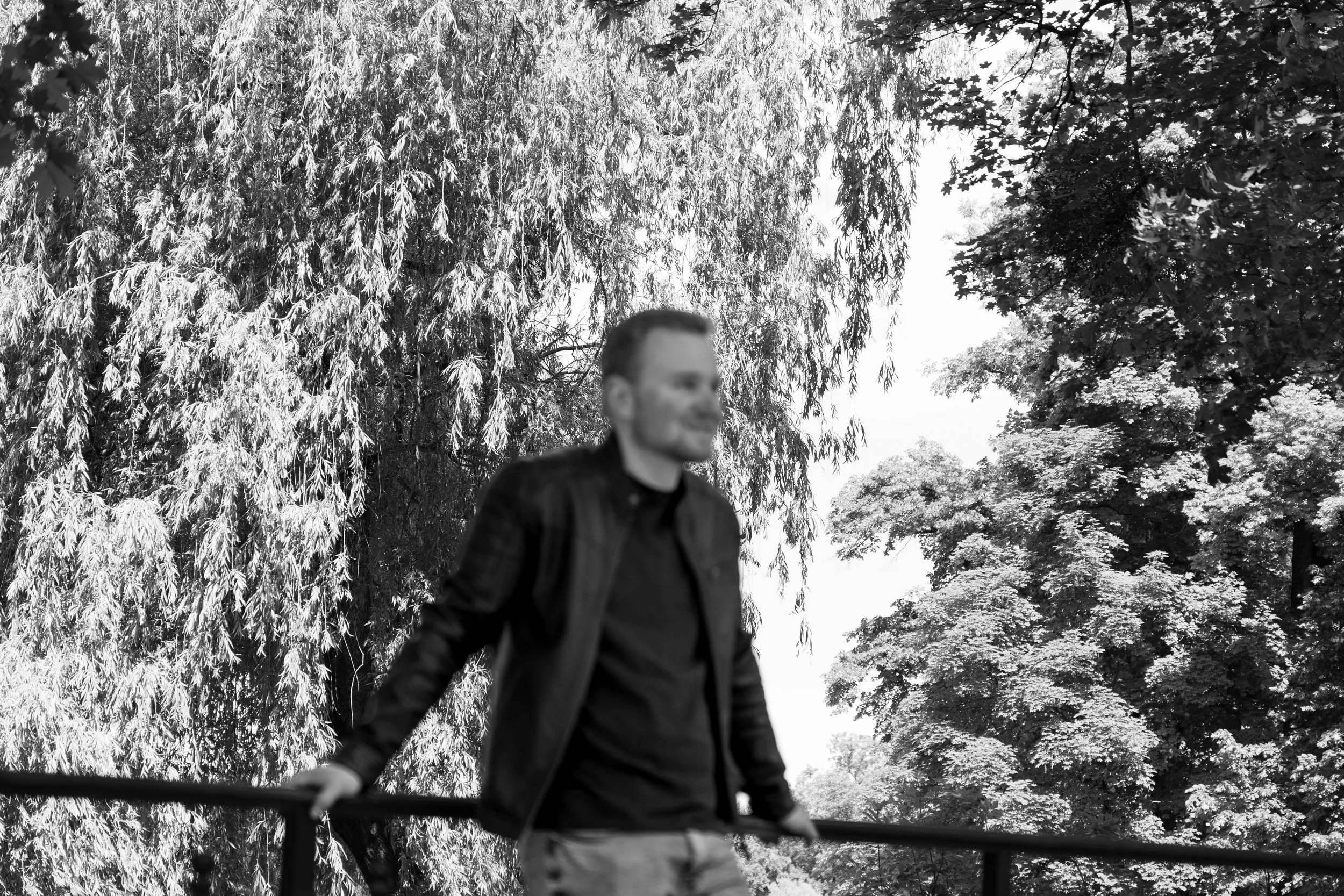 Ein lachender Mann mit dunkler Jacke und kurzen Haaren, der hinter einem Zaun läuft, im Hintergrund viele Bäume mit Blättern, schwarz-weiß Foto.