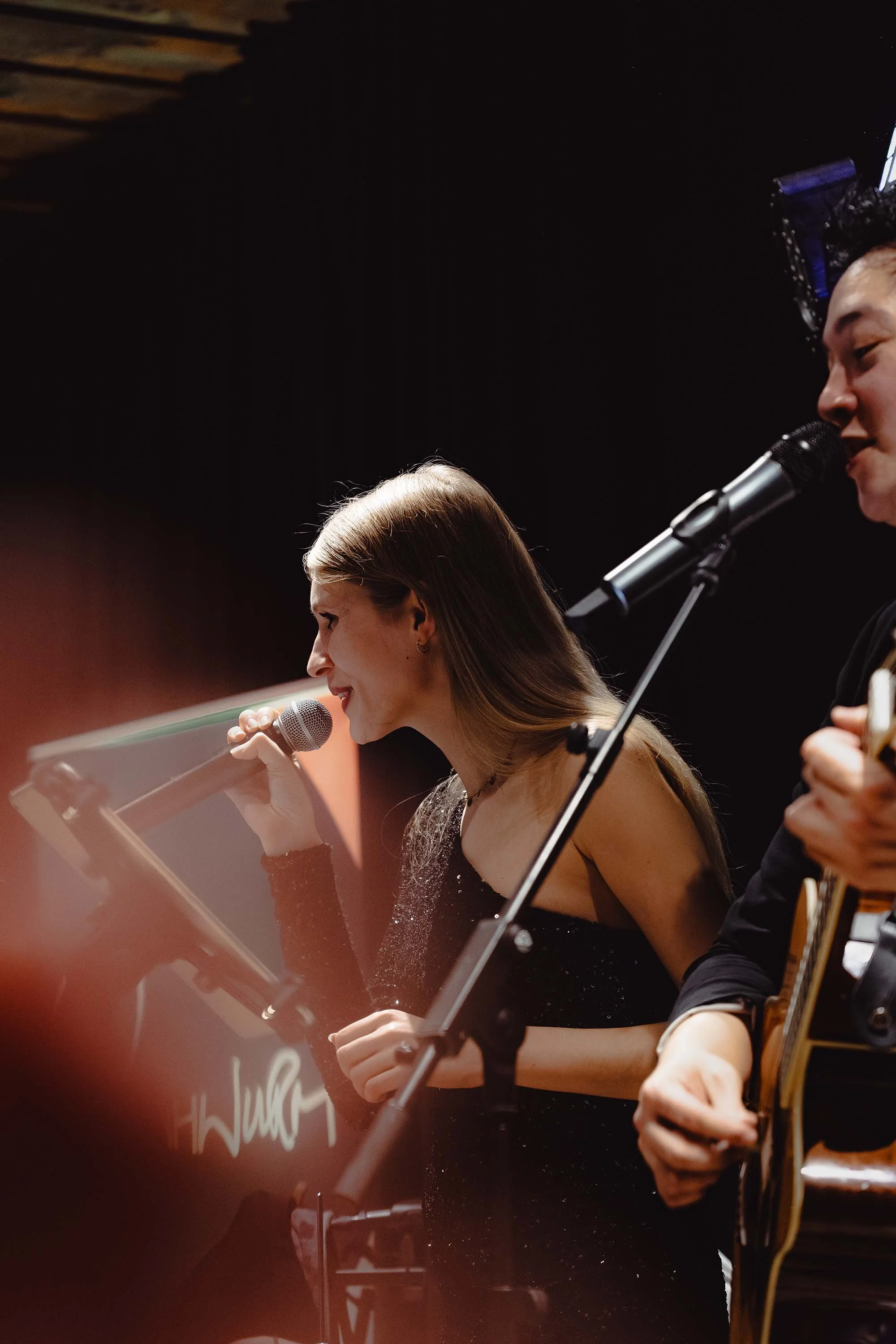 Eine Sängerin und ein Sänger auf der Bühne, die bei einem Musikauftritt singen, die Sängerin mit einem Mikrofon und der andere spielt Gitarre, vor einem schwarzen Hintergrund.