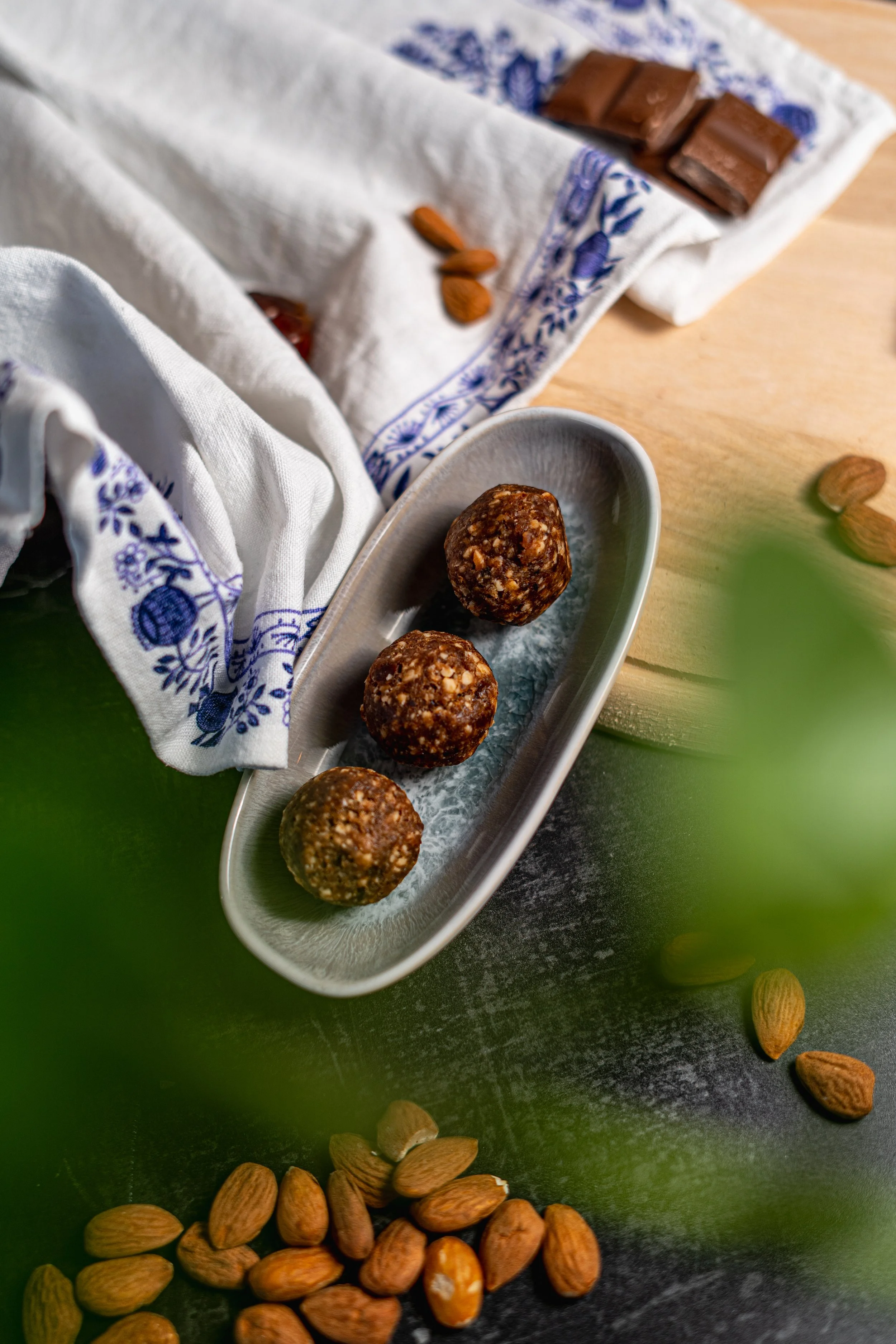 Drei Energieballs auf einer ovalen Schale, umgeben von Mandeln auf einer dunklen Tischfläche, mit einer dekorativen weinblauen weißen Stoffe und einem raschen Stoff im Hintergrund.