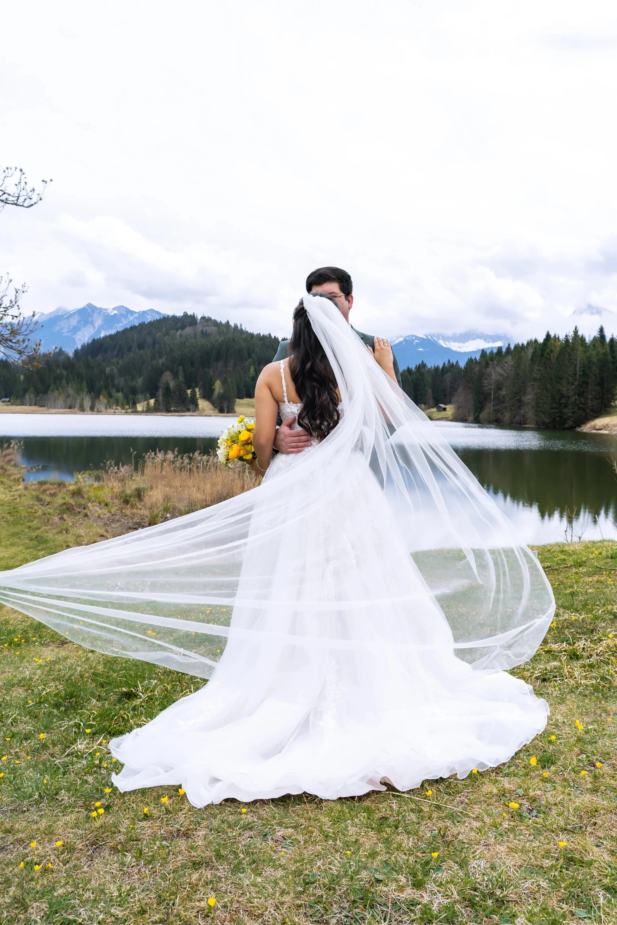 Ein Brautpaar steht am Wasser in einer bergigen Landschaft mit bewölktem Himmel. Die Braut trägt ein weißes Kleid mit langem Schleier, und der Bräutigam trägt einen dunklen Anzug. Die Braut hält einen Blumenstrauß in der Hand.