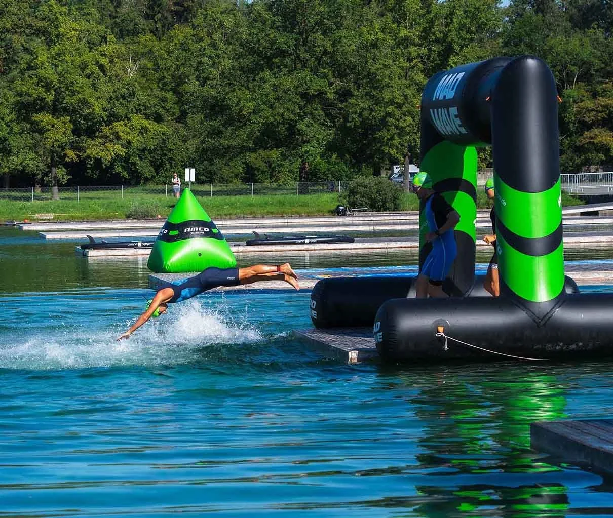 Ein Schwimmer springt ins Wasser während eines Wettkampfs, umgeben von einer Wasserbahn und Startblock, mit mehreren Personen im Wasser und am Ufer, im Hintergrund Bäume und Hohenzäune.