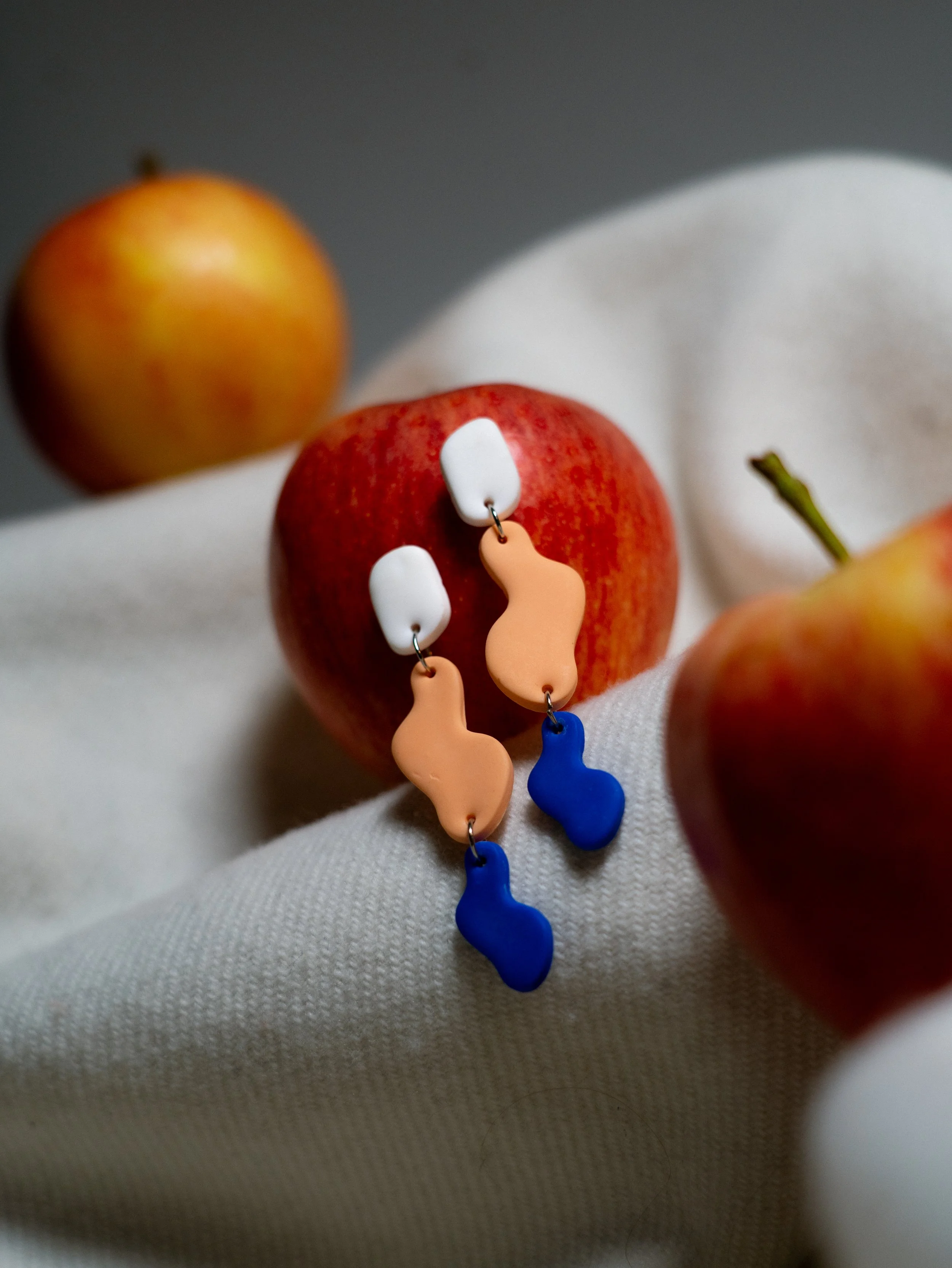 Nahaufnahme eines roten Apfels mit bunten Ohrringen; im Hintergrund gibt es einen weiteren, unscharfen Apfel und eine weiße Stofffläche.