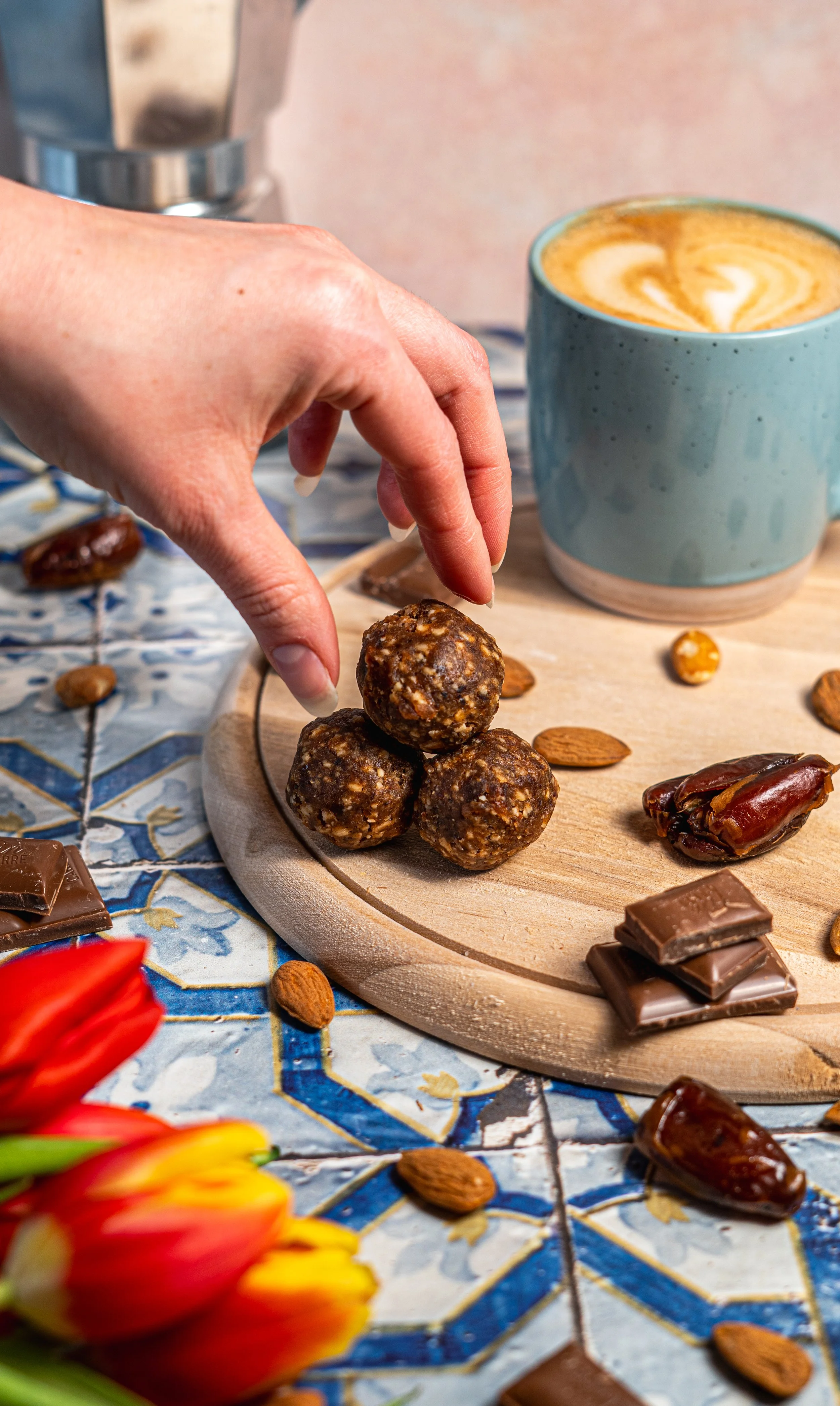 Ein Hand greift nach mehreren Energyballs, die auf einem runden Holzbrett liegen. Neben dem Brett befindet sich eine blau-gekachelte Oberfläche mit Schokoladenstücken, Mandeln, Datteln und einer Tasse mit Kaffee mit Milchschaum.