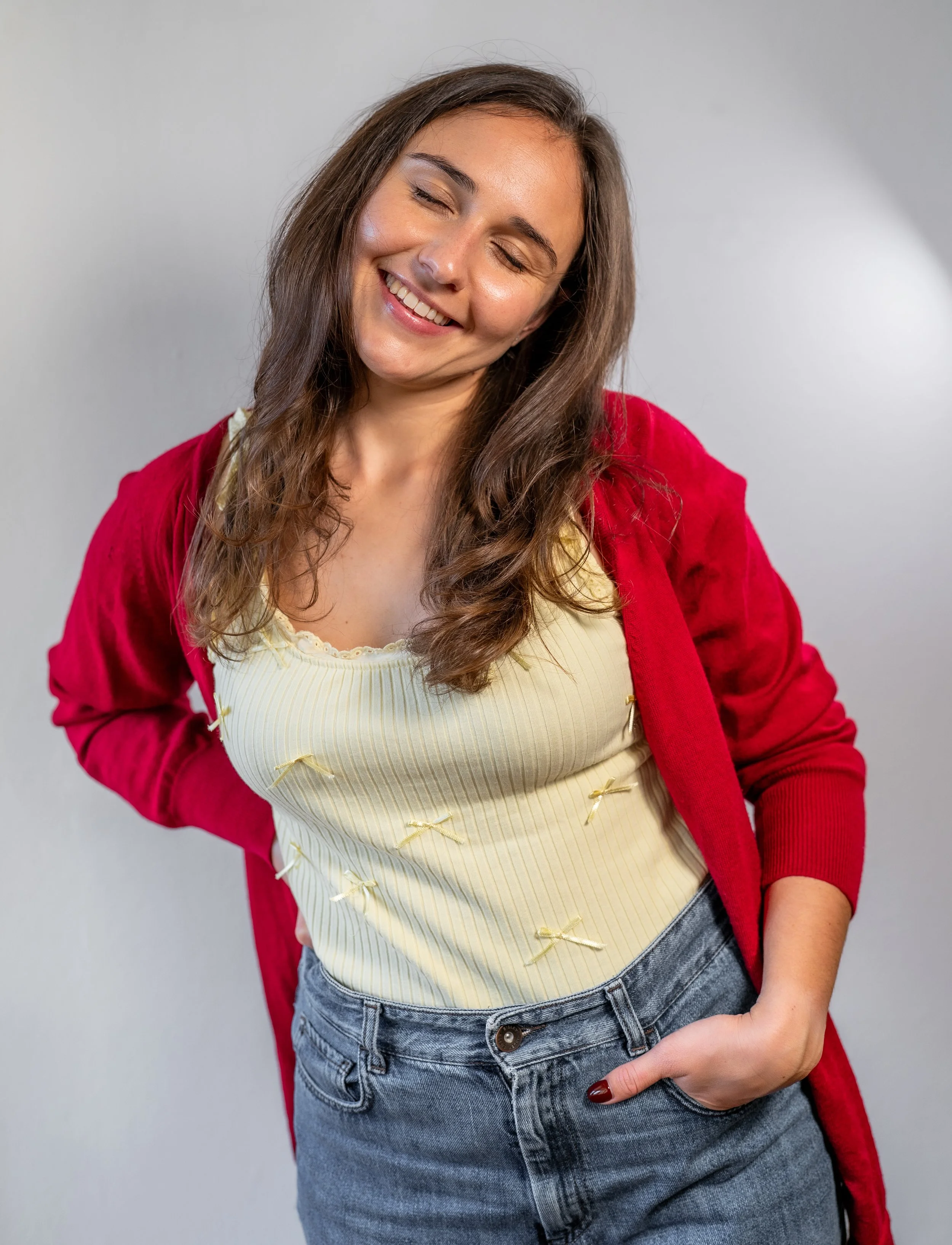 Junge Frau trägt rote Jacke und gelbe Bluse mit Rüschen, mit ihren Augen geschlossen und lächelnd, in Jeans, vor einem grauen Hintergrund.