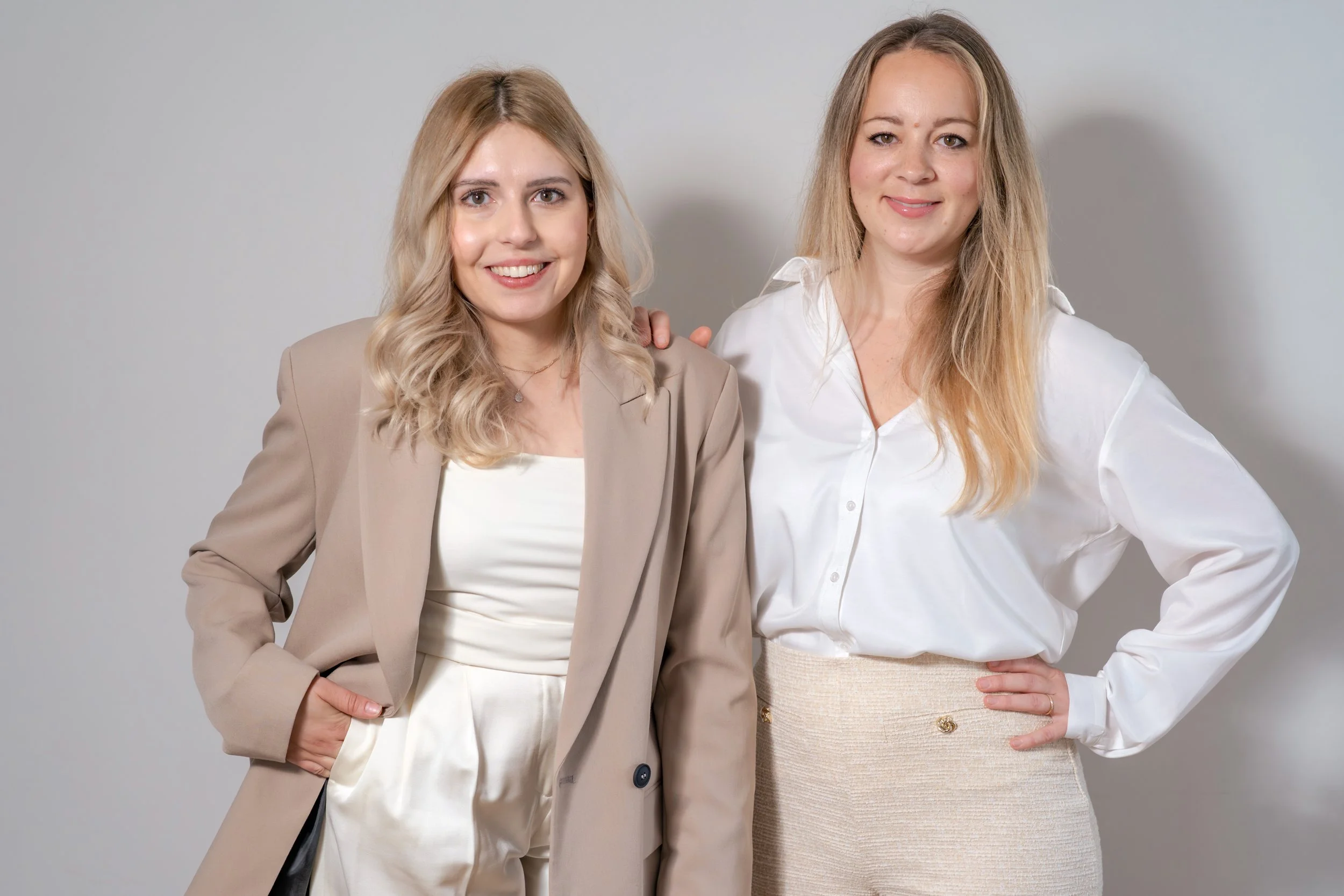 Zwei Frauen stehen nebeneinander vor einem grauen Hintergrund, lächeln und schauen in die Kamera, eine trägt einen beigen Blazer und die andere einen weißen Blazer.