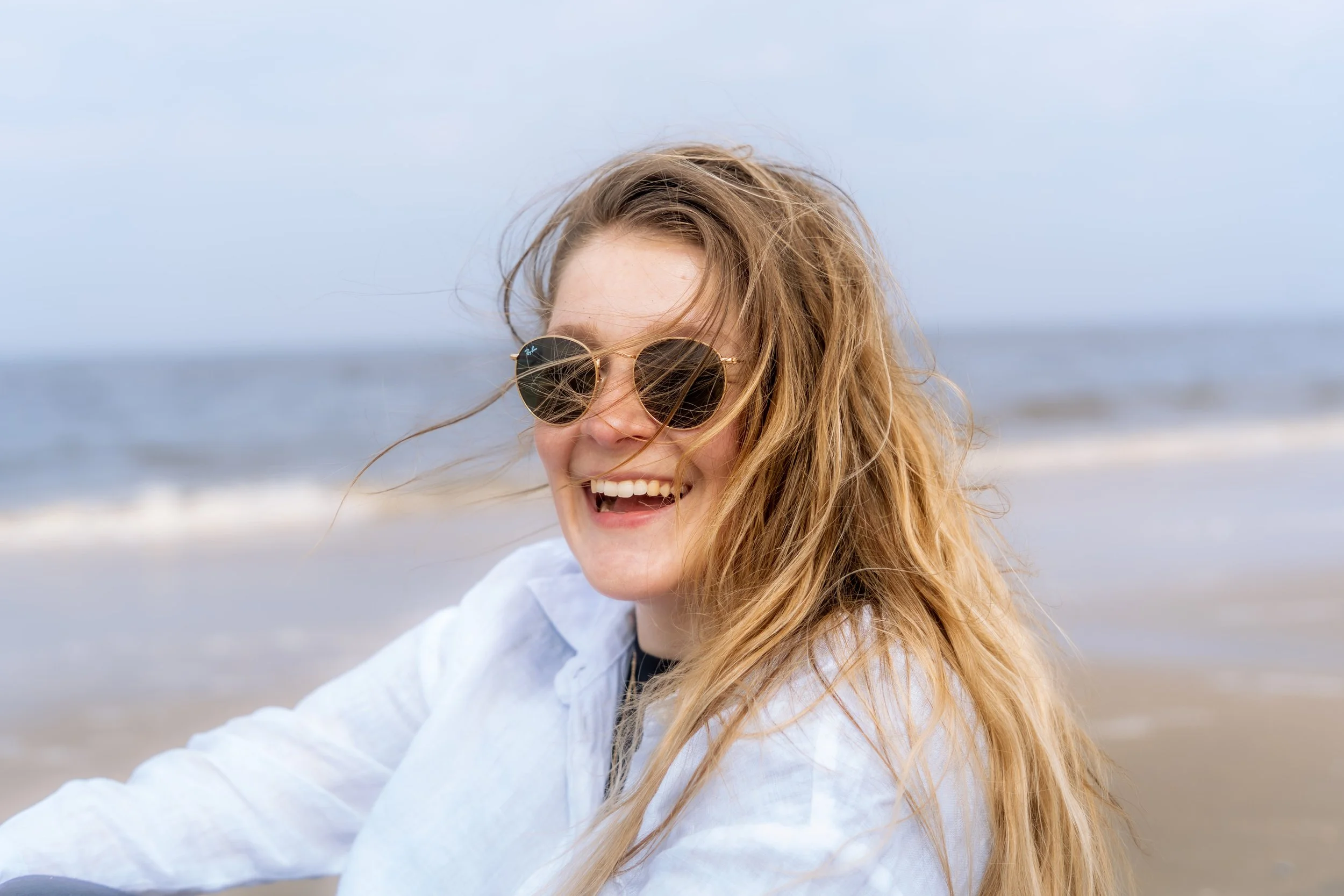 Junge Frau mit lockigem Haar trägt runde Sonnenbrille und lacht am Strand.
