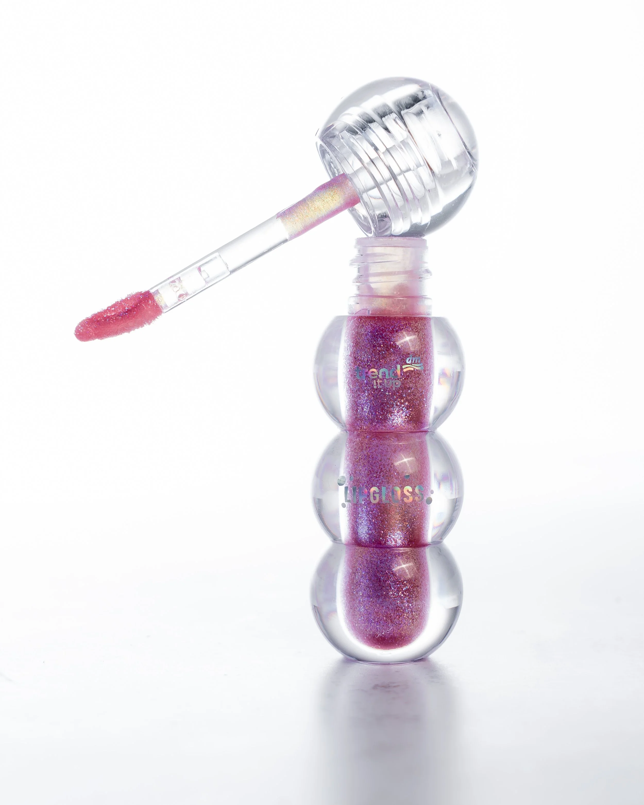 Lippgloss mit geöffnetem Deckel und Pinsel, der den pinkfarbenen, glitzernden Lippgloss zeigt, vor weißem Hintergrund und reflektiert auf der Oberfläche.