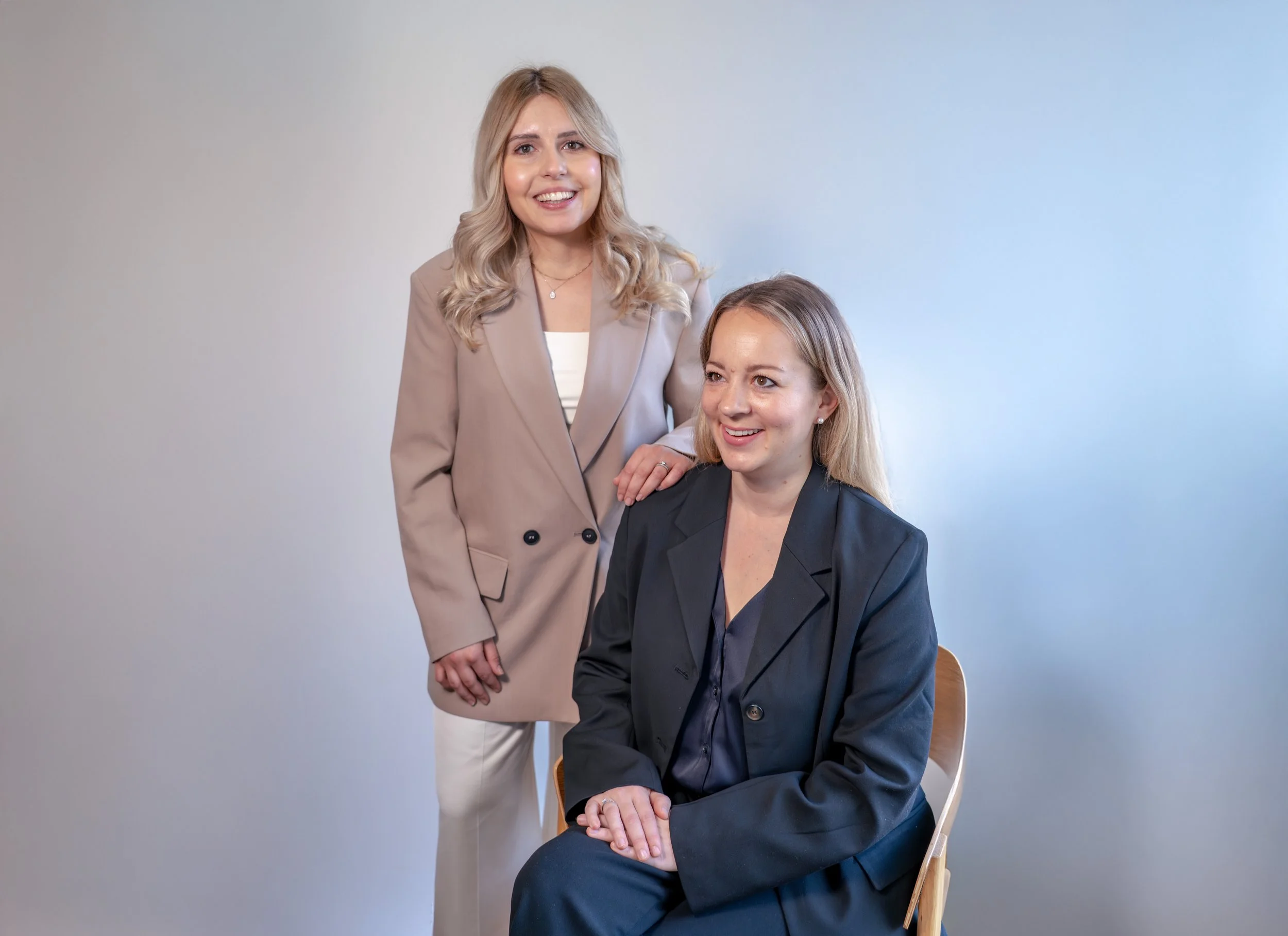 Zwei Frauen in einem professionellen Umfeld, eine steht, die andere sitzt auf einem Stuhl, beide lächeln. Helles, schlichtes Hintergrundlevel.