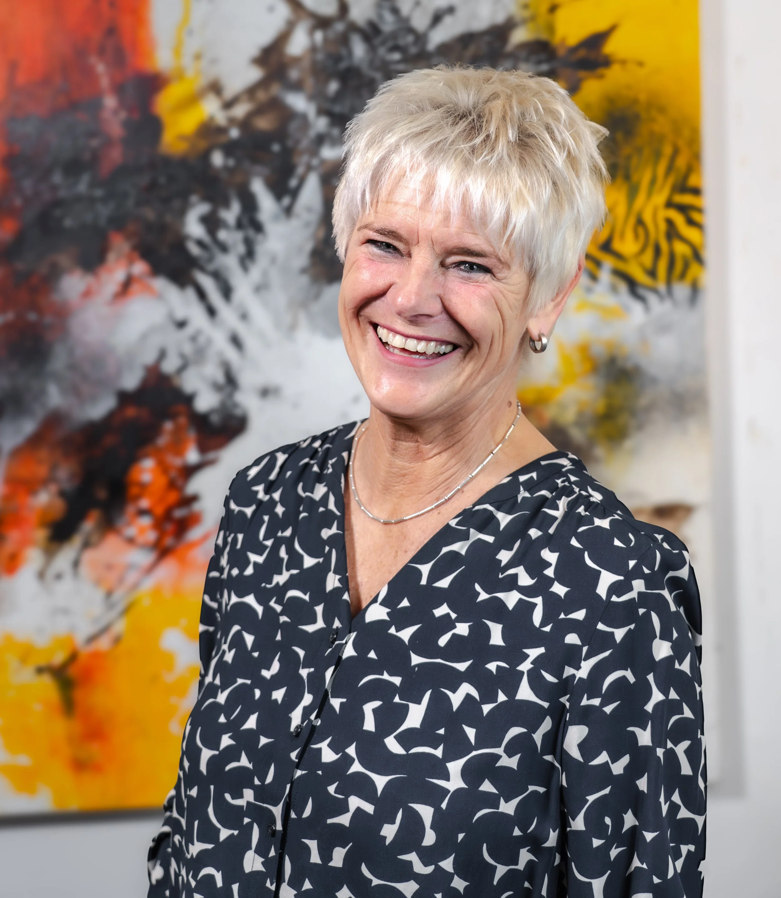 Eine lachende Frau mit kurzen, blond gefärbten Haaren trägt ein schwarzes Kleid mit weißem Muster und steht vor einer abstrakten bunten Kunstinstallation.
