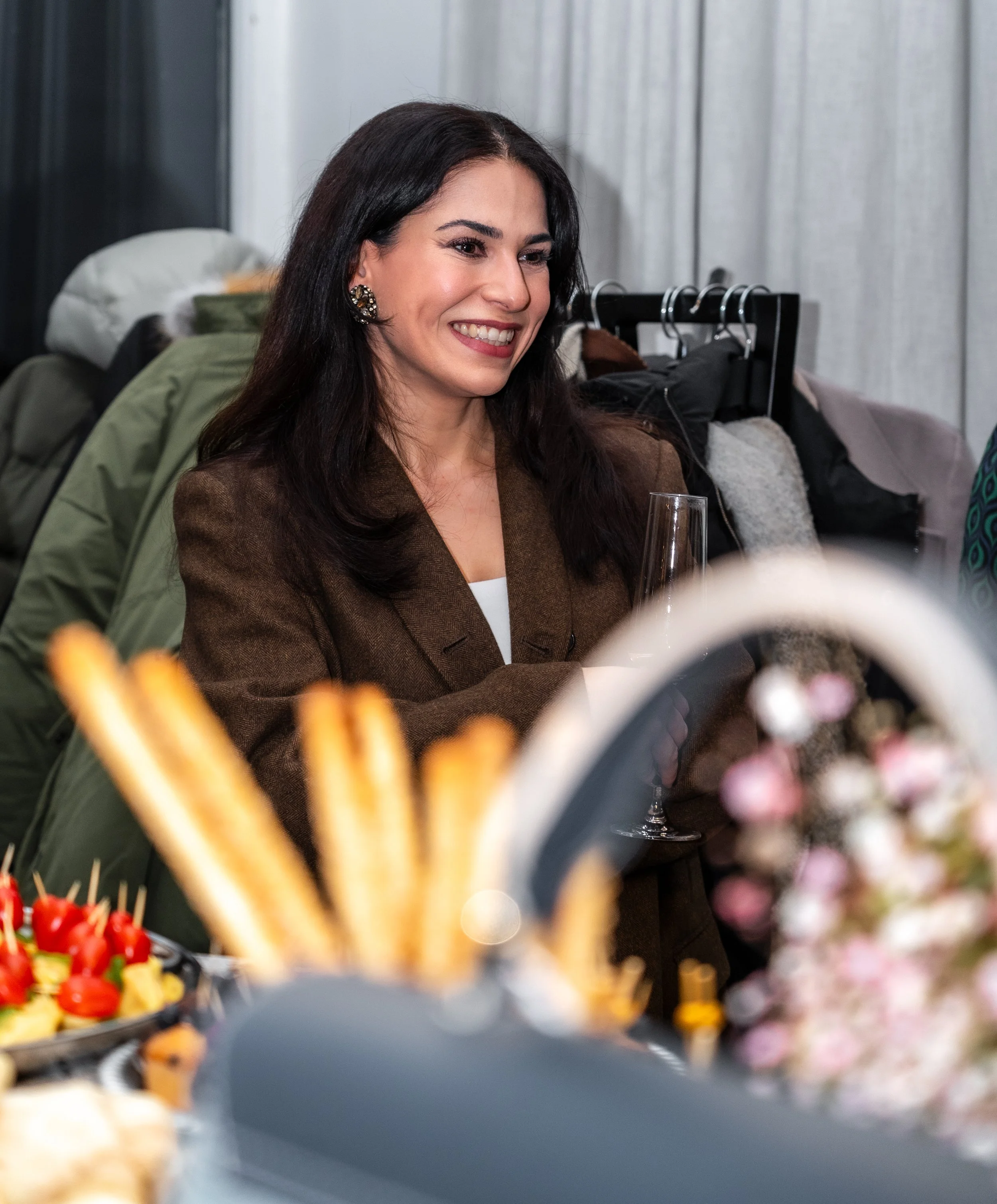 Lächelnde Frau mit dunklen Haaren in einem braunen Mantel bei einer Party, im Hintergrund Kleidung auf Kleiderständern, vorderer Bereich mit Fingerfood und Blumen.