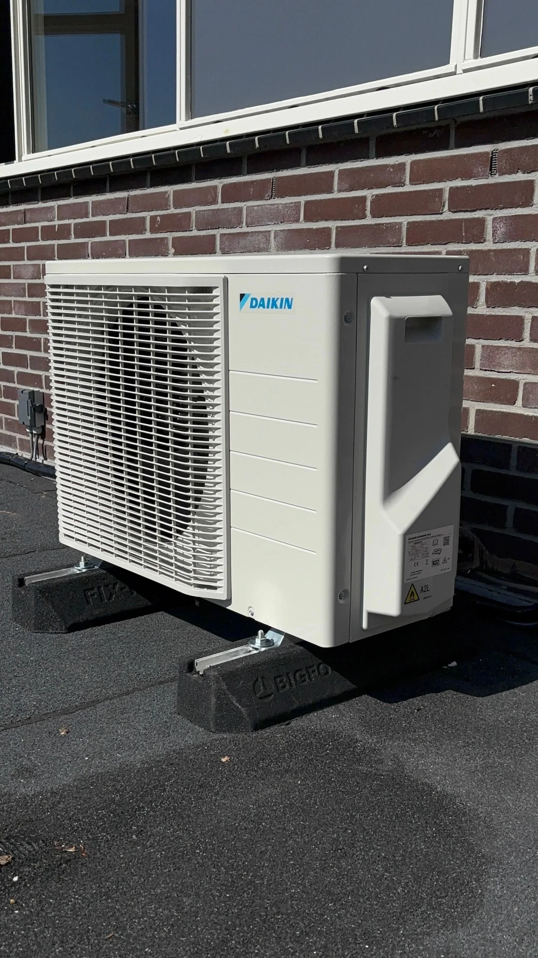 Een buitenunit van een airconditioningsysteem van het merk Daikin, geïnstalleerd op een dak met een asfaltoppervlak voor een gebouw met een bakstenen muur.