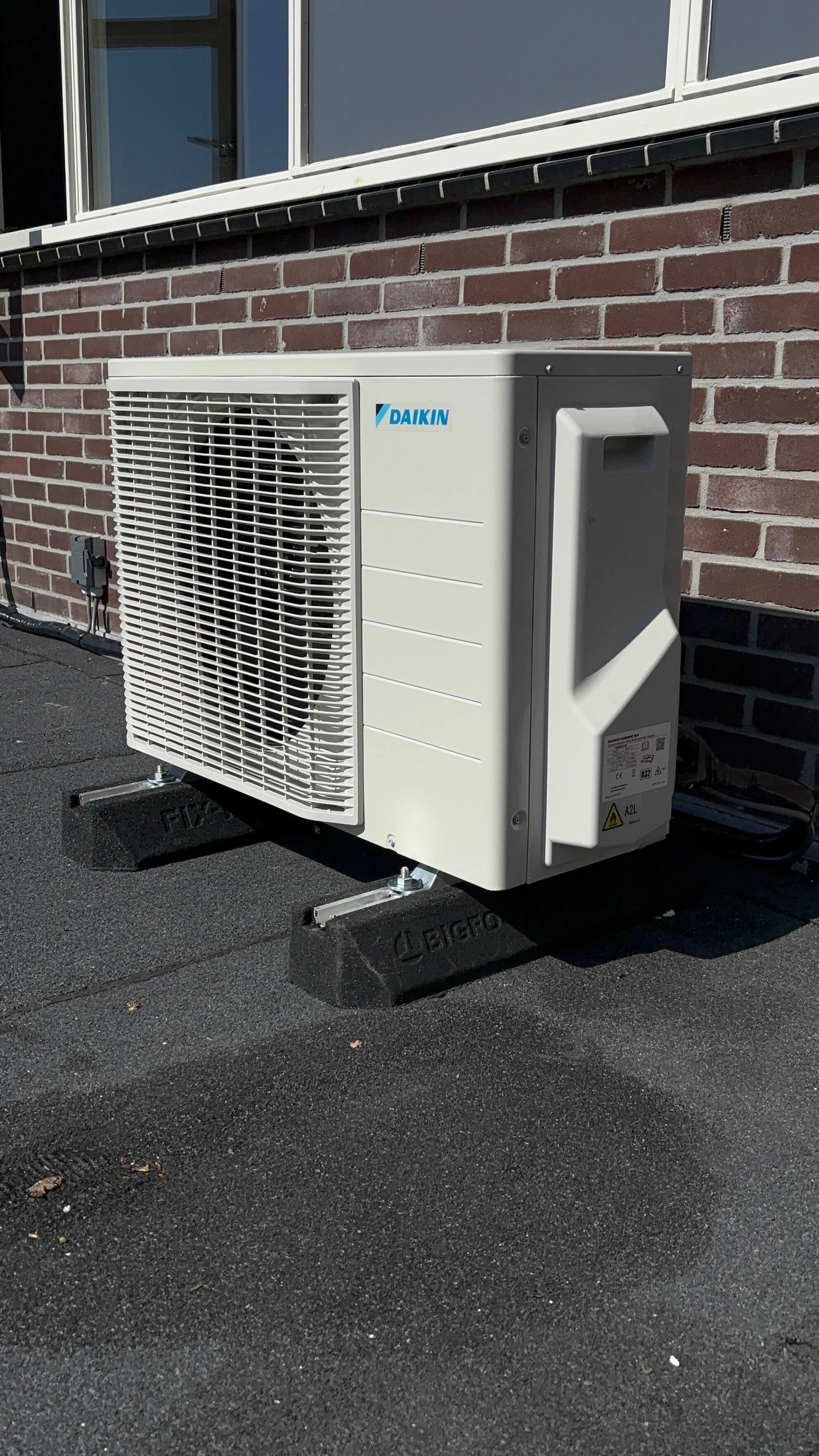 Een buitenunit van een airconditioningsysteem van het merk Daikin, geïnstalleerd op een zwart dakvlak voor een gevel met bakstenen muur.