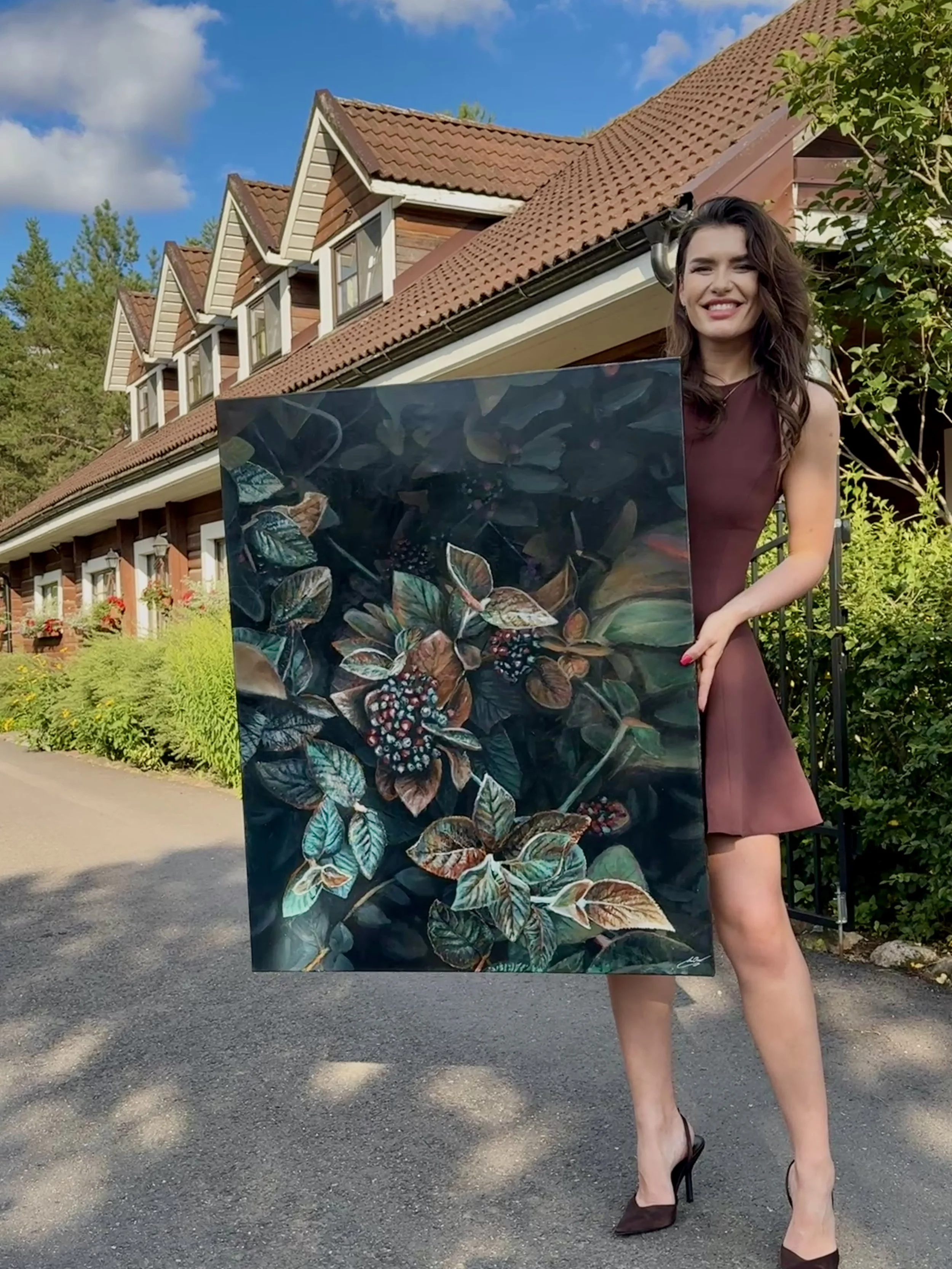 A woman with wavy brown hair and a burgundy dress standing outdoors in front of a house with a red tiled roof and dormer windows, holding a large painting of leafy plants with berries.