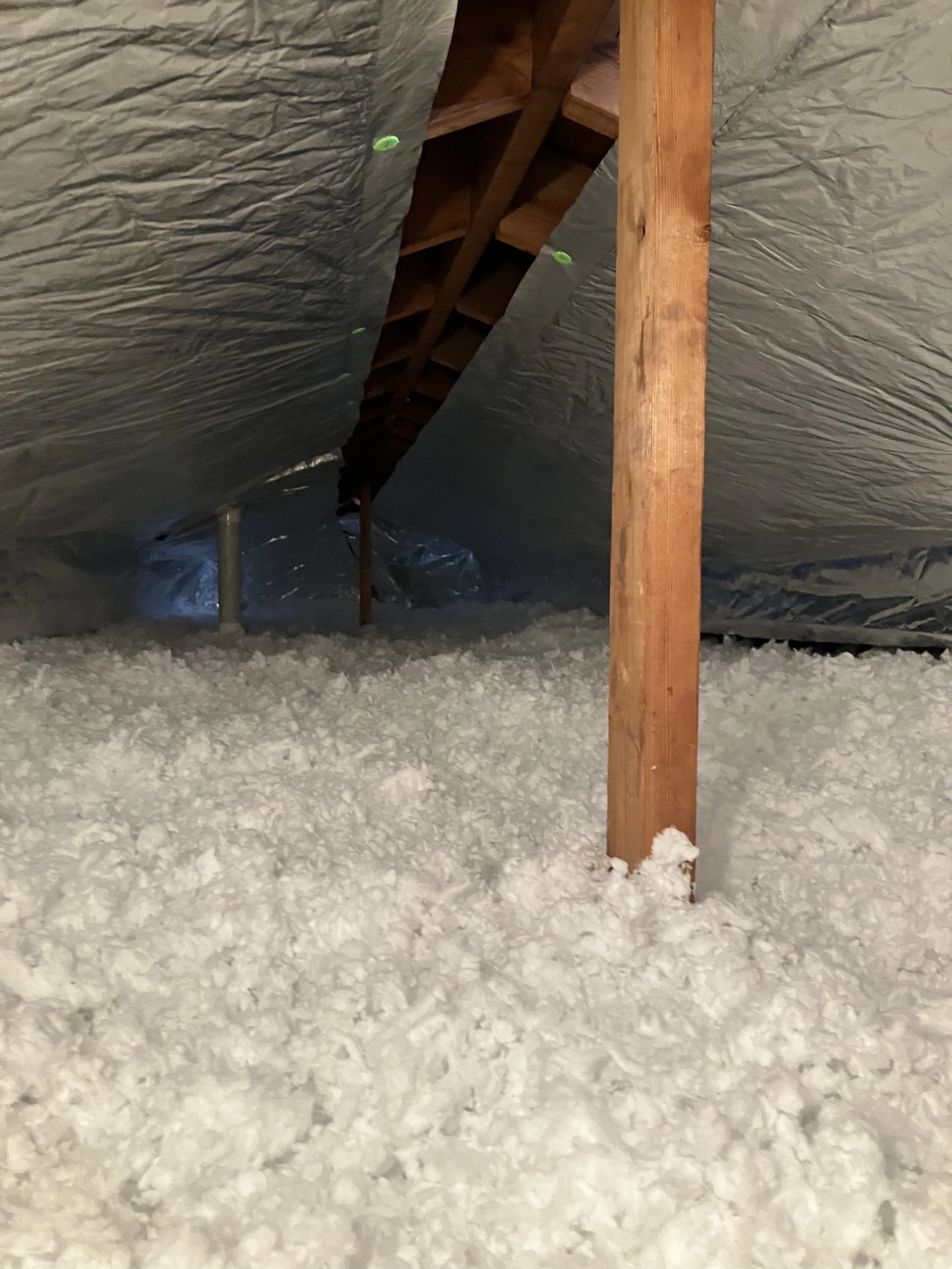 Attic space with foam insulation, wooden support beams, and a wooden roof structure visible. The floor is covered with some insulation material or snow.