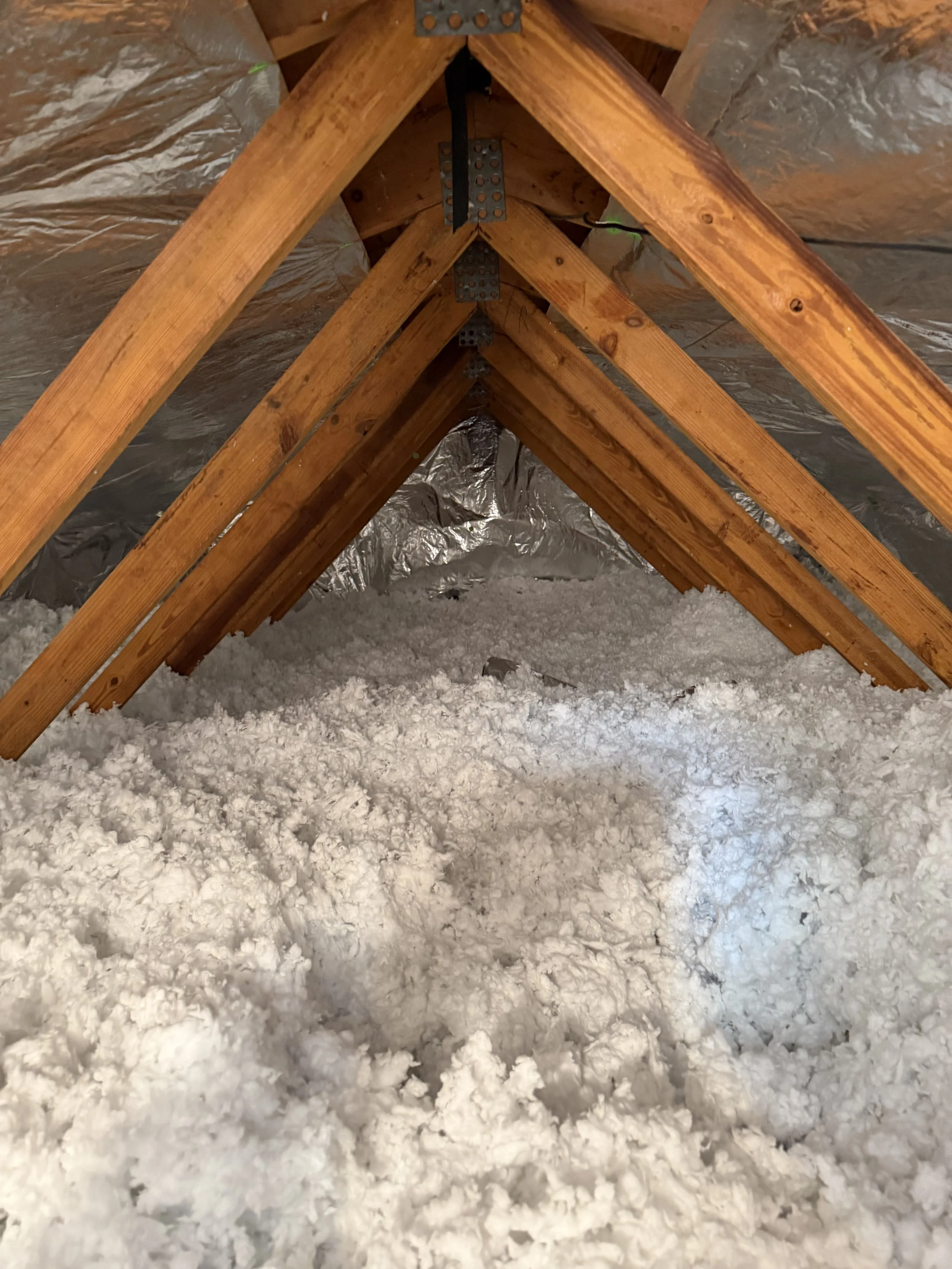 Attic space with wood framing, insulation, and a layer of white spray foam insulation on the floor.