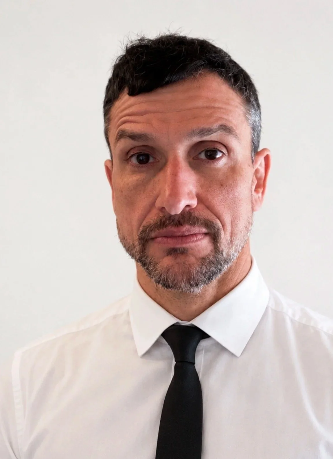 A man with short dark hair, some gray, and a beard, wearing a white shirt and black tie, looking directly at the camera with a neutral expression, against a plain light background.