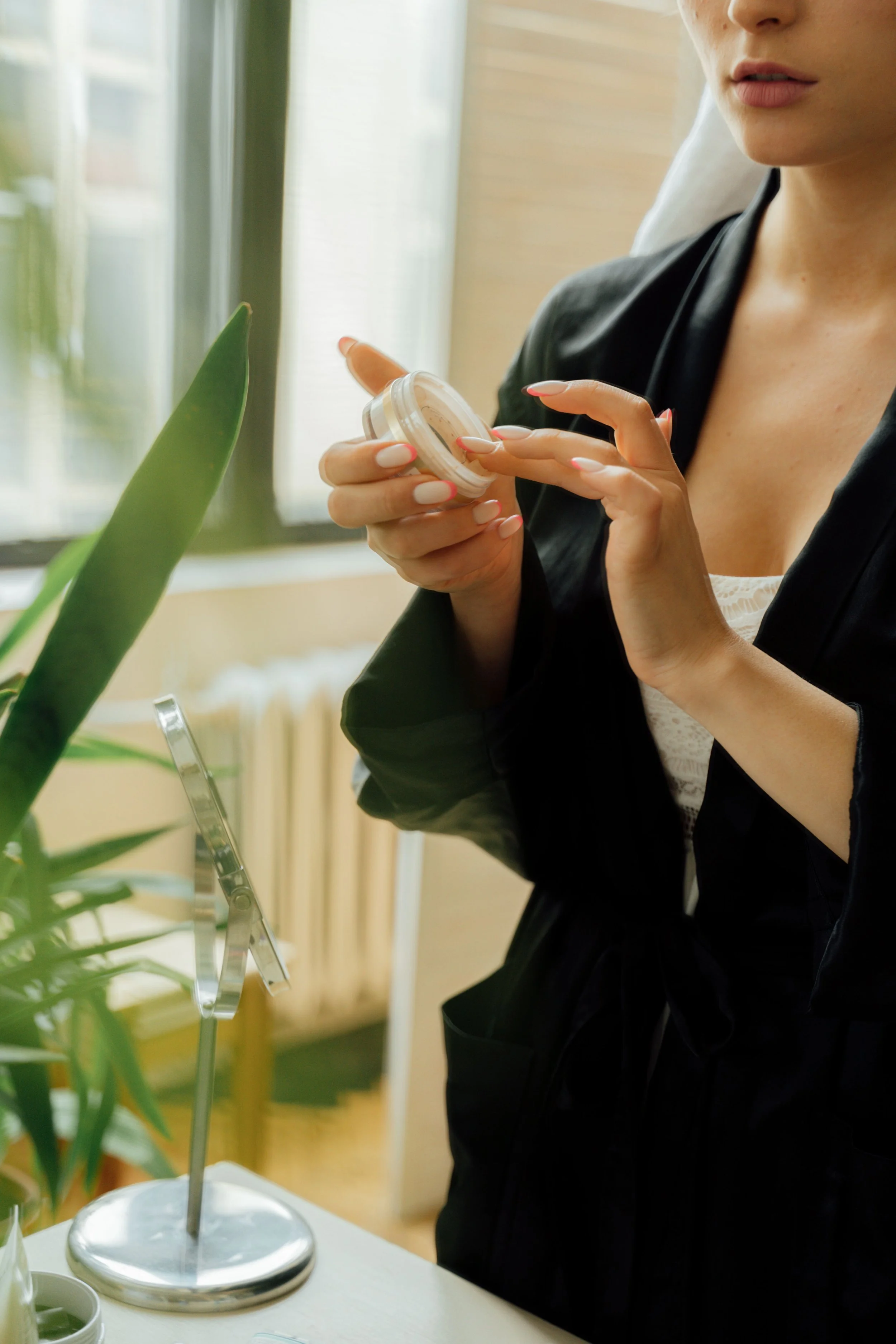 A woman with light skin and painted nails is holding a container near her face. She is wearing a black satin top. The background includes a window with sunlight and a plant with broad green leaves.