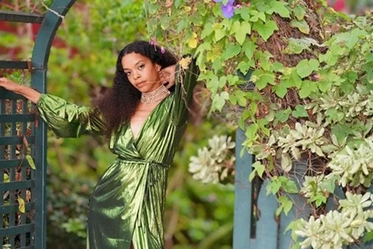 A young woman in a shiny green dress standing in a garden next to a metal gate and surrounded by green plants and flowers.Photographer, for hire, Oceanside, Carlsbad, San Diego, Orange County, Fashion, Portraits, Moments, Lifestyle, Events, Branding.