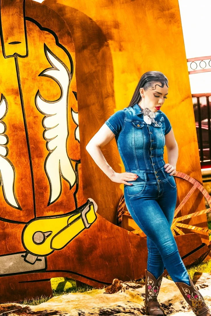 A woman in a denim outfit standing outdoors in front of a western-themed backdrop with a painted design of a fork and coal miner pickaxe.