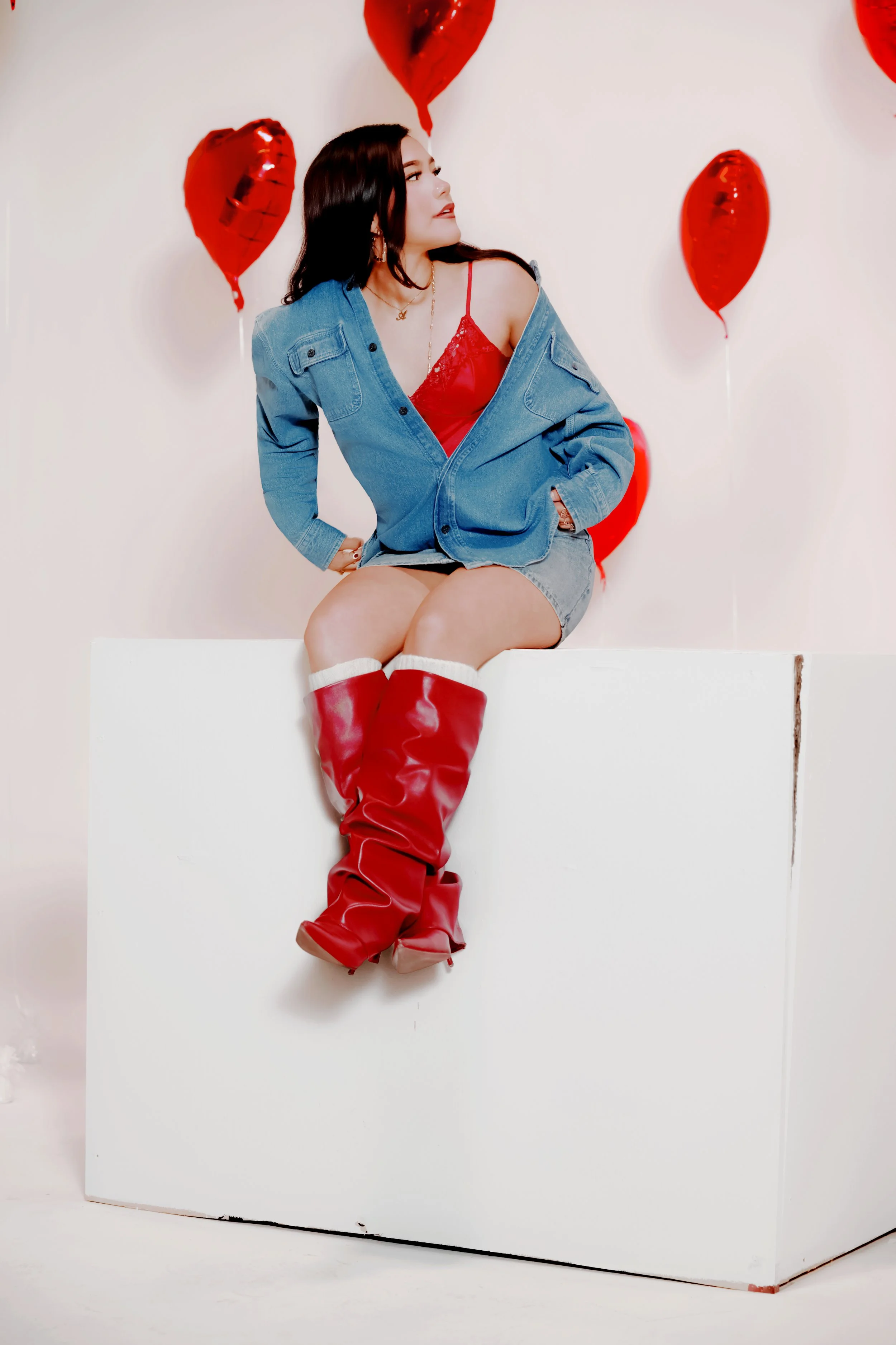 A young woman with dark hair sitting on a white block, wearing a red top, denim jacket off her shoulders, and red thigh-high boots, surrounded by red heart-shaped balloons against a plain white background.Studio, Photographer, Hire, Book .