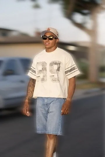 Photographer, for hire, Oceanside, Carlsbad, San Diego, Orange County, Fashion, Portraits, Moments, Lifestyle, Events, Branding.Man wearing sunglasses, a beige cap, a white vintage-style t-shirt with number 92, and denim shorts, walking on a street.
