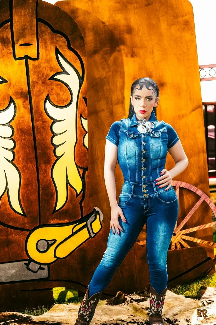 A woman standing in front of a wooden backdrop with a painted cowboy boot and hat. She is wearing a denim jumpsuit, cowboy boots, and has styled curly hair with bright makeup.