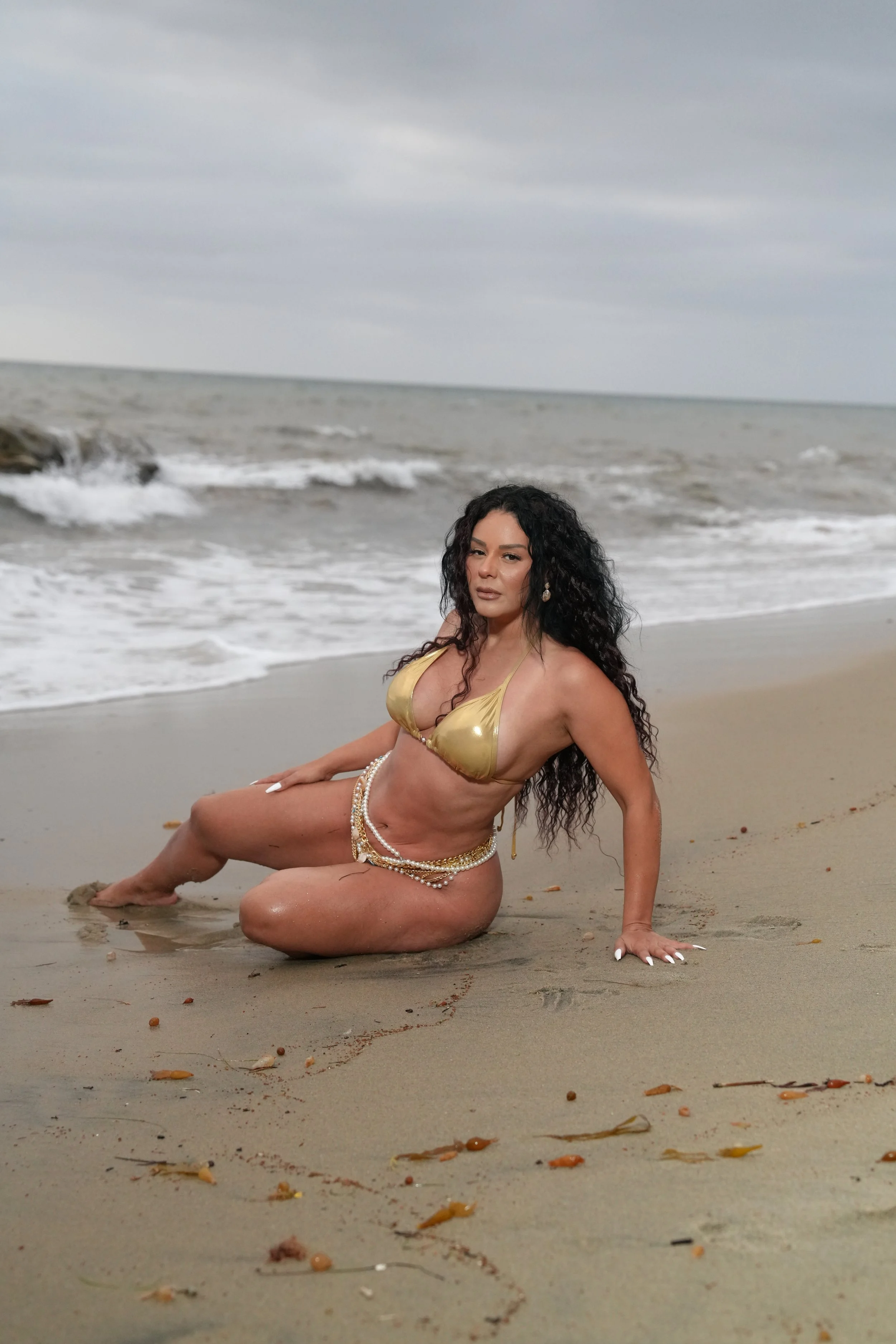 A woman with long black curly hair, wearing a gold bikini top and pearl accessories, is reclining on a sandy beach with ocean waves and cloudy sky in the background.