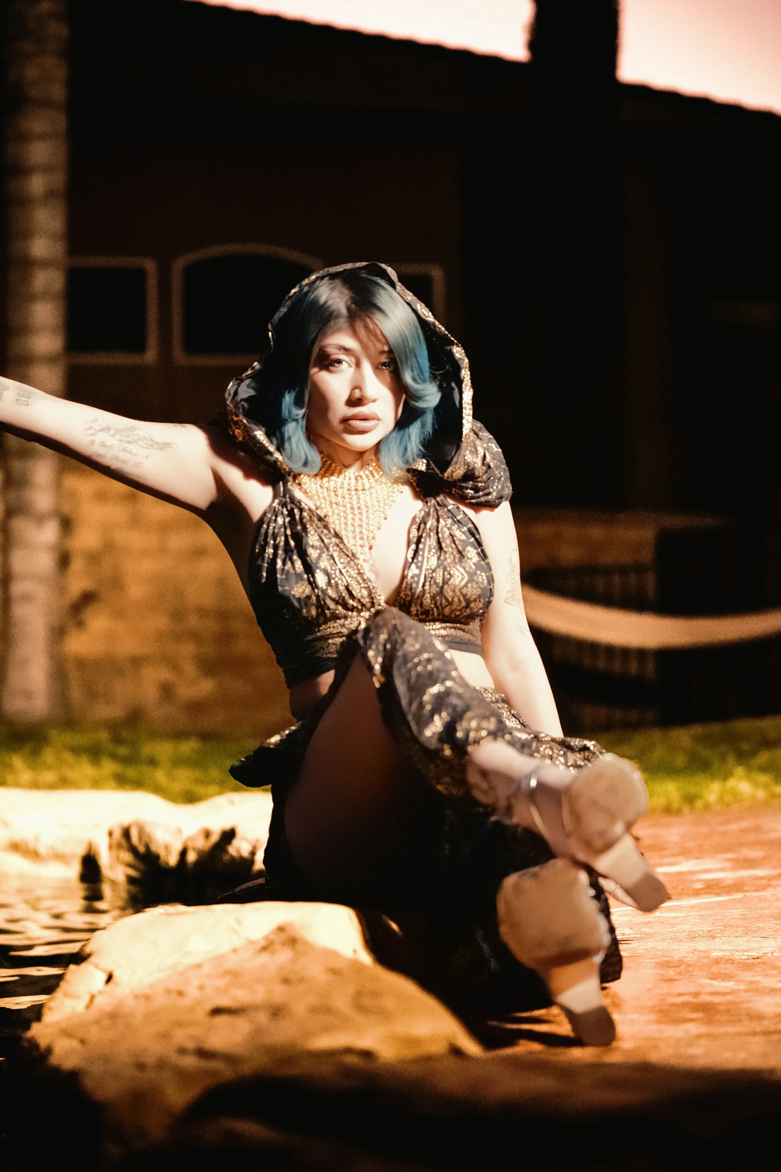 A woman with blue hair and tattoos sitting by a body of water during sunset, wearing a patterned black and gold hooded outfit and boots.Photographer, for hire, Oceanside, Carlsbad, San Diego, Orange County, Fashion, Portraits, Moments, Lifestyle.