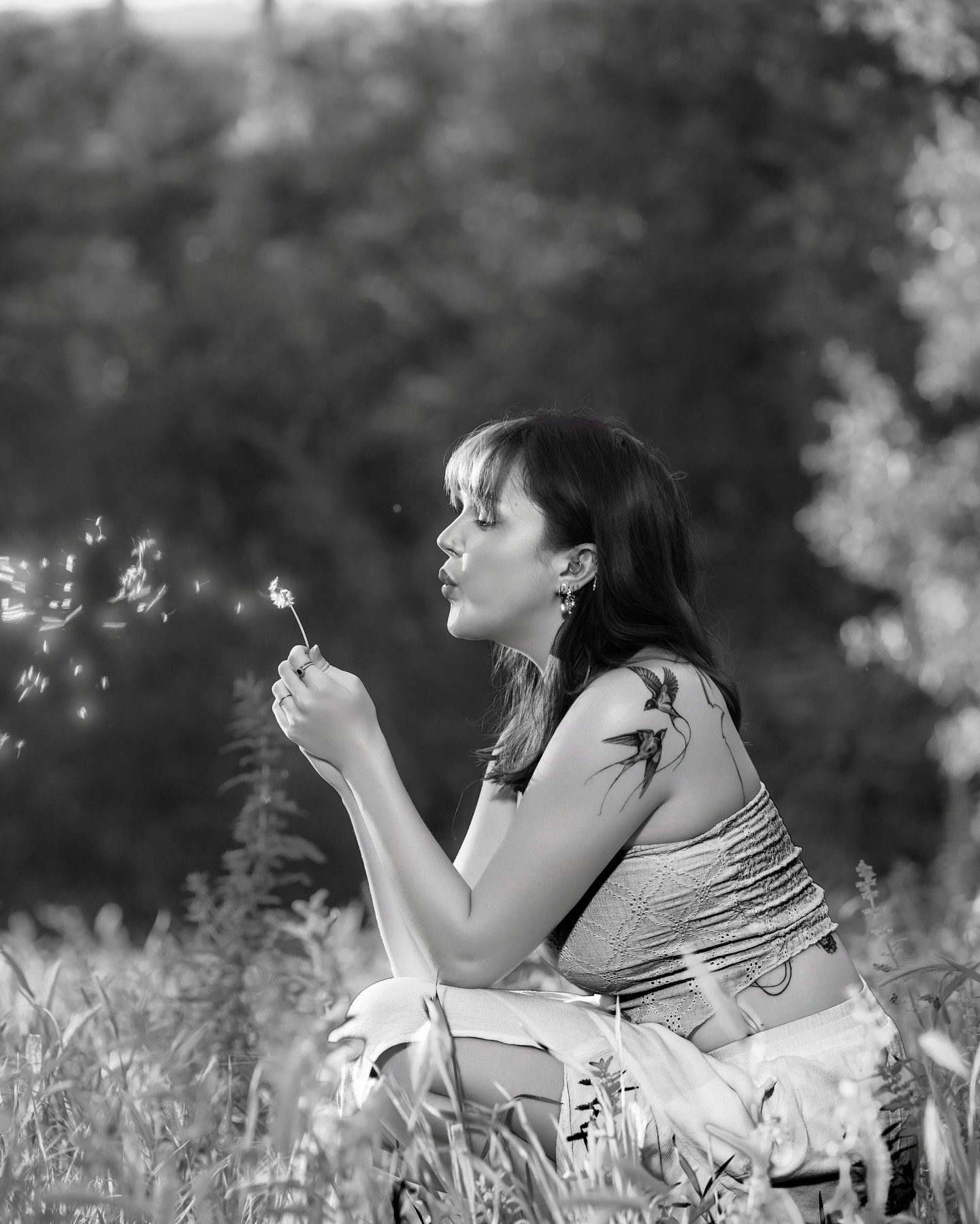 A young woman with dark hair, tattoos, and earrings, sitting outdoors in a grassy area, blowing on a dandelion in a moment of serene contemplation, with blurred trees in the background.
San Diego editorial photographer
High-end portraiture services