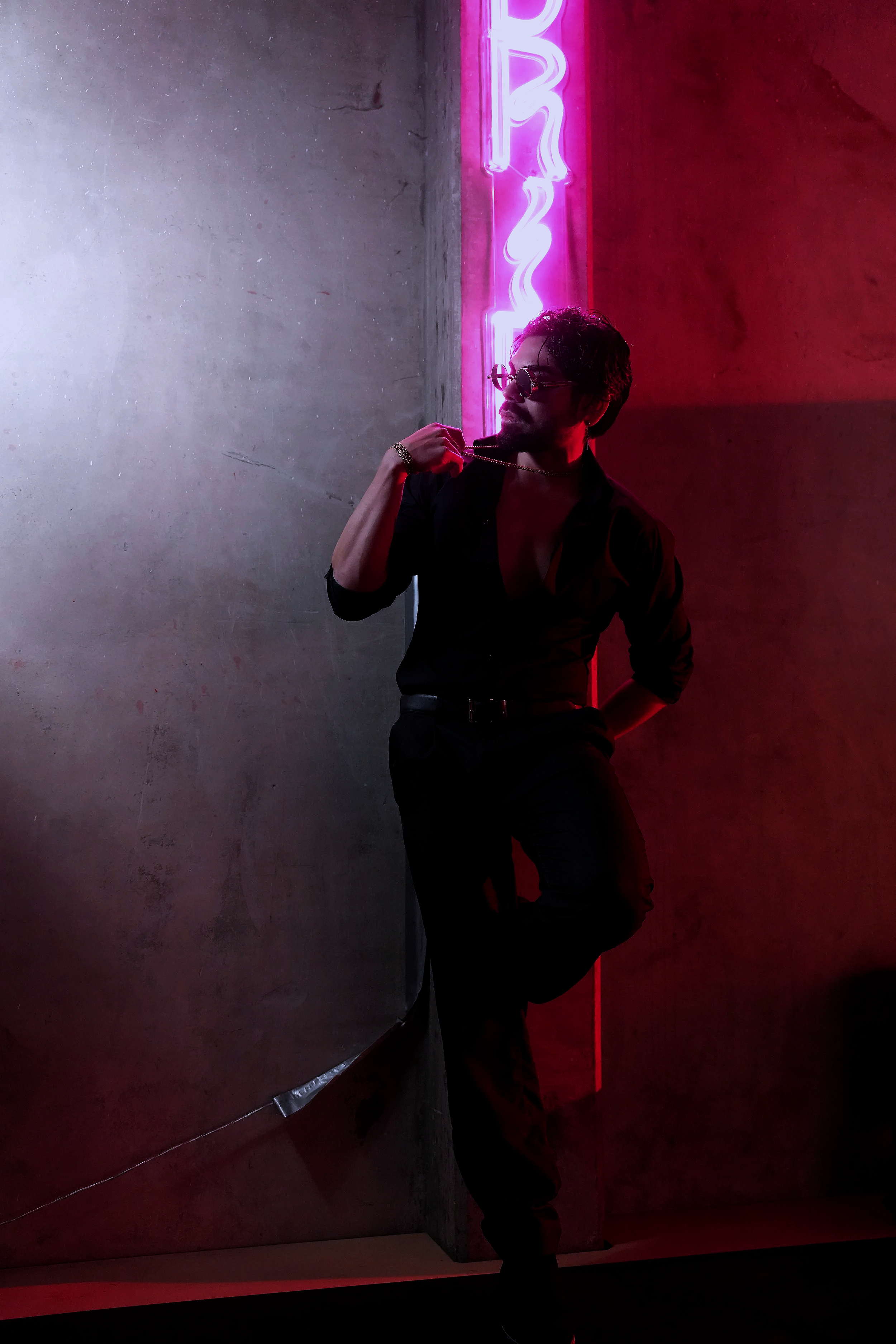 A man with curly hair and sunglasses, wearing a black shirt and pants, is standing with one hand on his chest and the other behind his back in front of a concrete wall with neon pink and purple signage in the background.