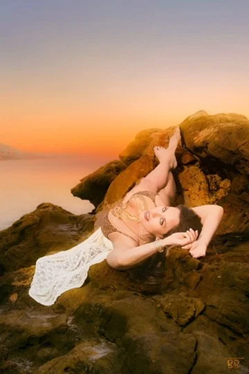 PhotographerPhotographer, for hire, Oceanside, Carlsbad, San Diego, Orange County, Fashion, Portraits, Moments, Lifestyle, Events, Branding.A woman in a beige lace dress lying on large rocks at sunset, posing with her arm behind her head and smiling.