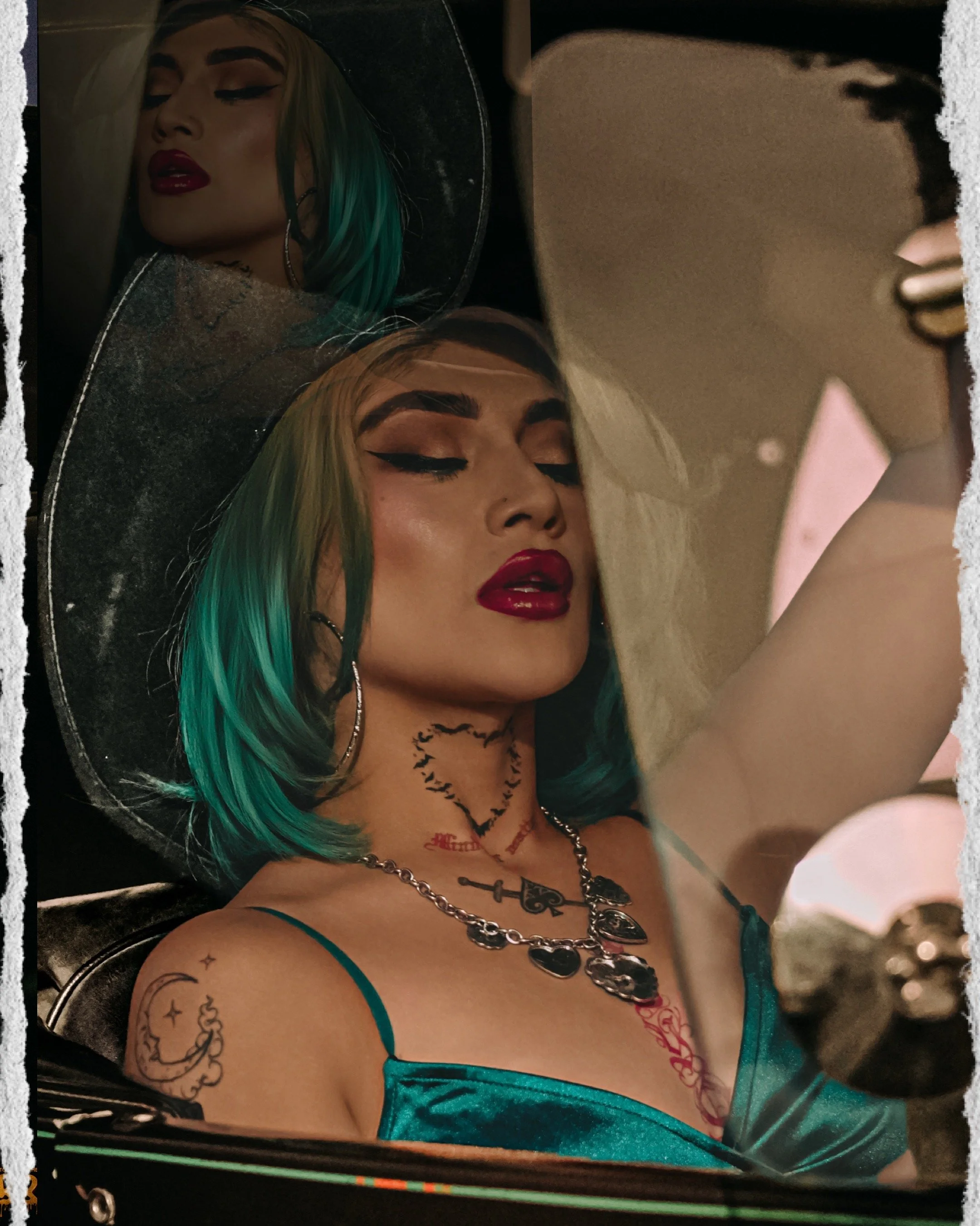 A woman with blonde hair that has turquoise tips, wearing makeup with bold eyeliner and red lipstick, and jewelry including layered necklaces, is taking a selfie in a mirror. She has tattoos on her shoulder and chest and is wearing a teal-colored sat