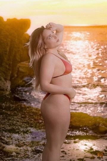 Photographer, for hire, Oceanside, Carlsbad, San Diego, Orange County, Fashion, Portraits, Moments, Lifestyle, Events, BrandingA woman in a red bikini standing on a beach at sunset, with her hand on her head and the ocean and rocks in the background.
